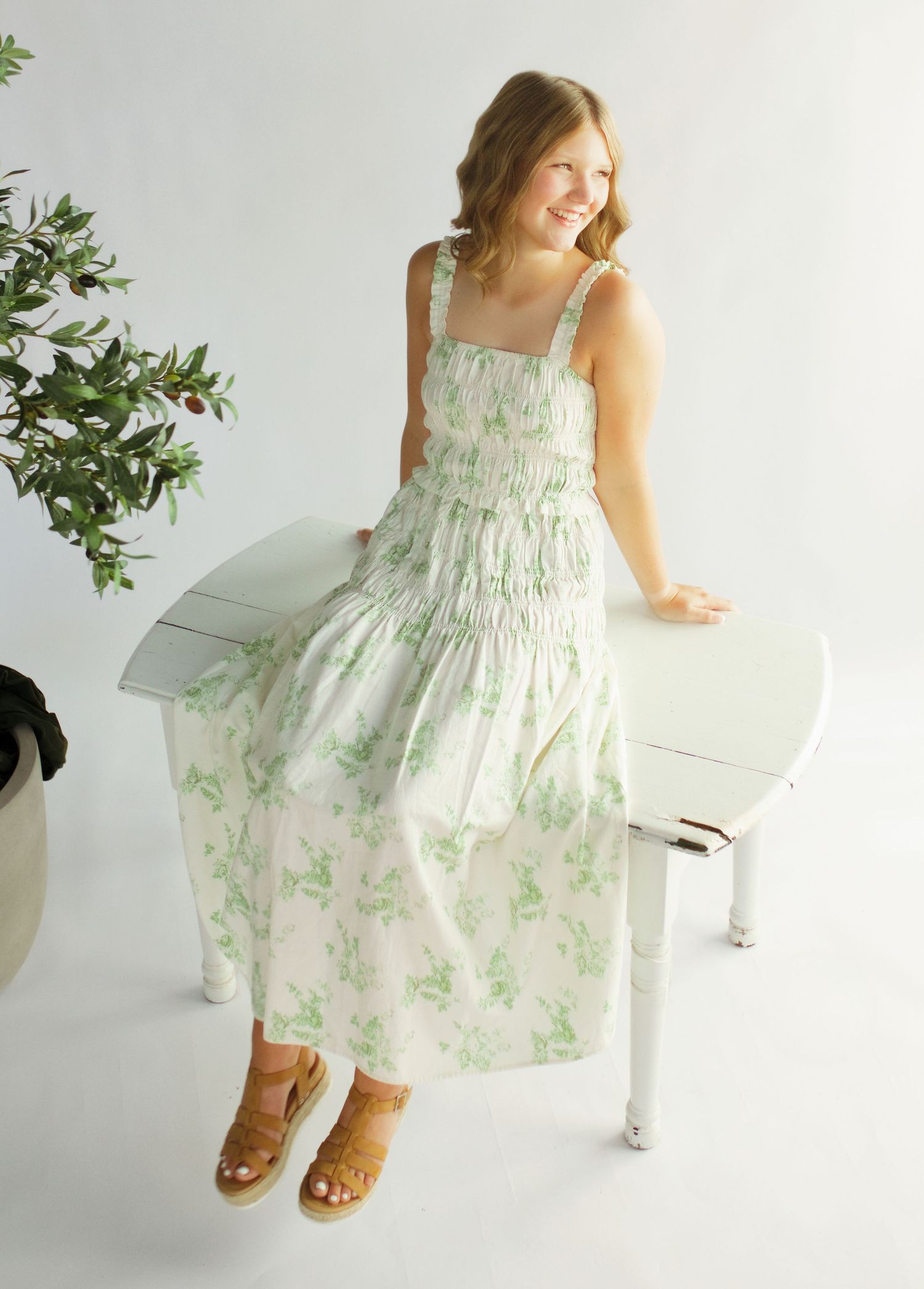 Woman wearing a white and green floral smocked maxi dress with tan sandals, posing near a white piano.