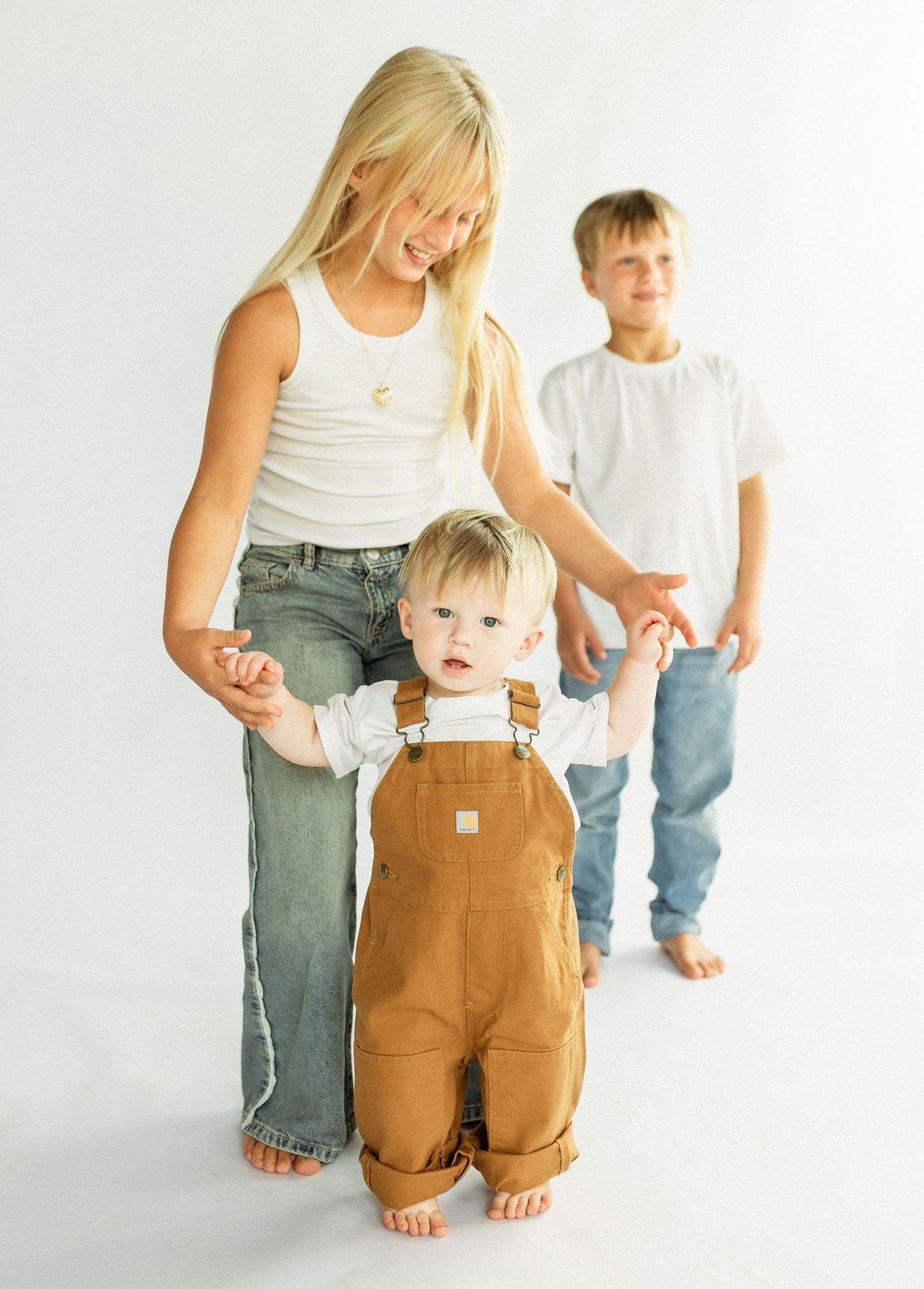Three children pose together, with a smiling blonde girl helping a toddler in tan overalls stand, while a boy watches.