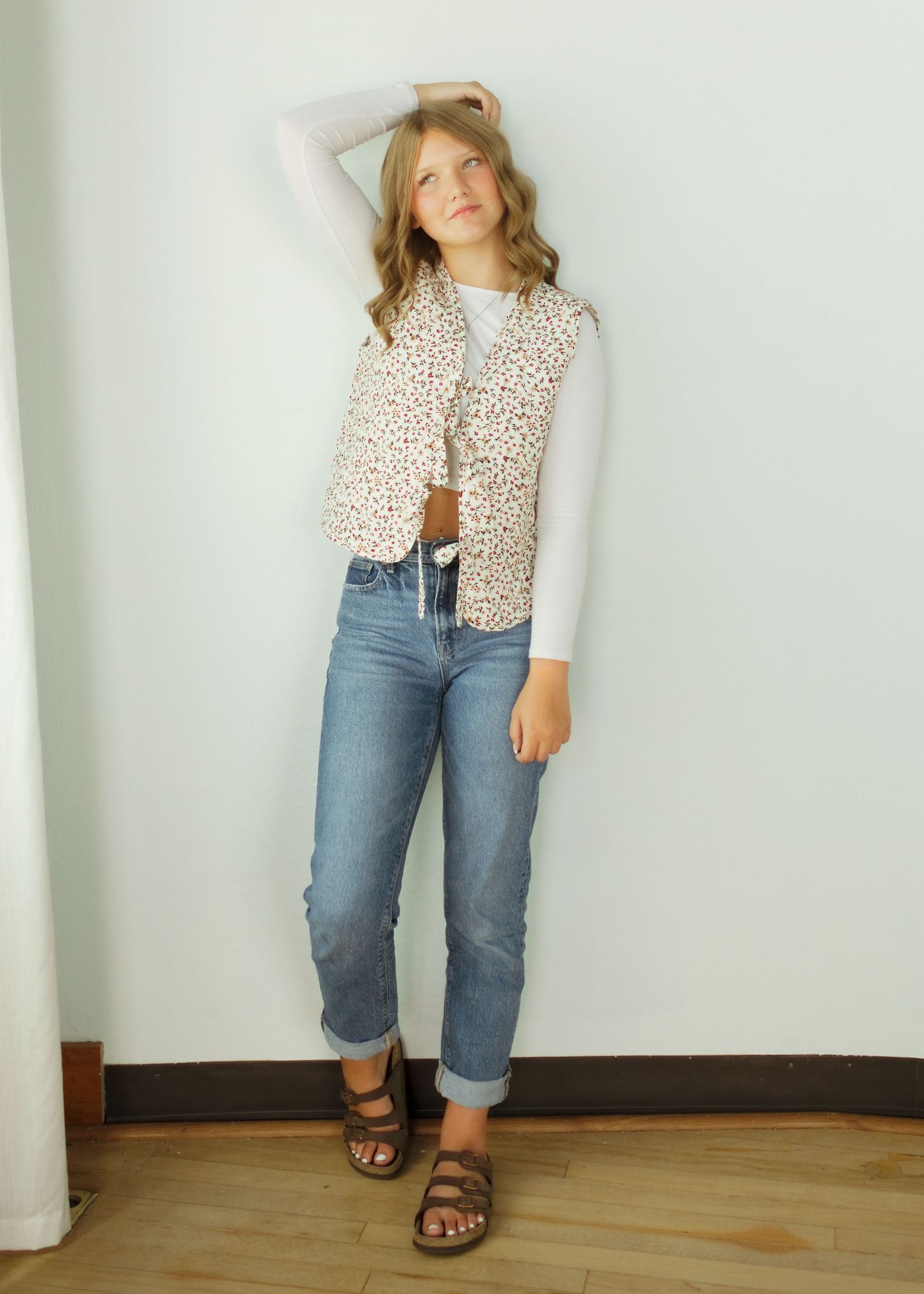 Teen girl wearing a floral vest over a white long-sleeve top with rolled cuff jeans and brown sandals.