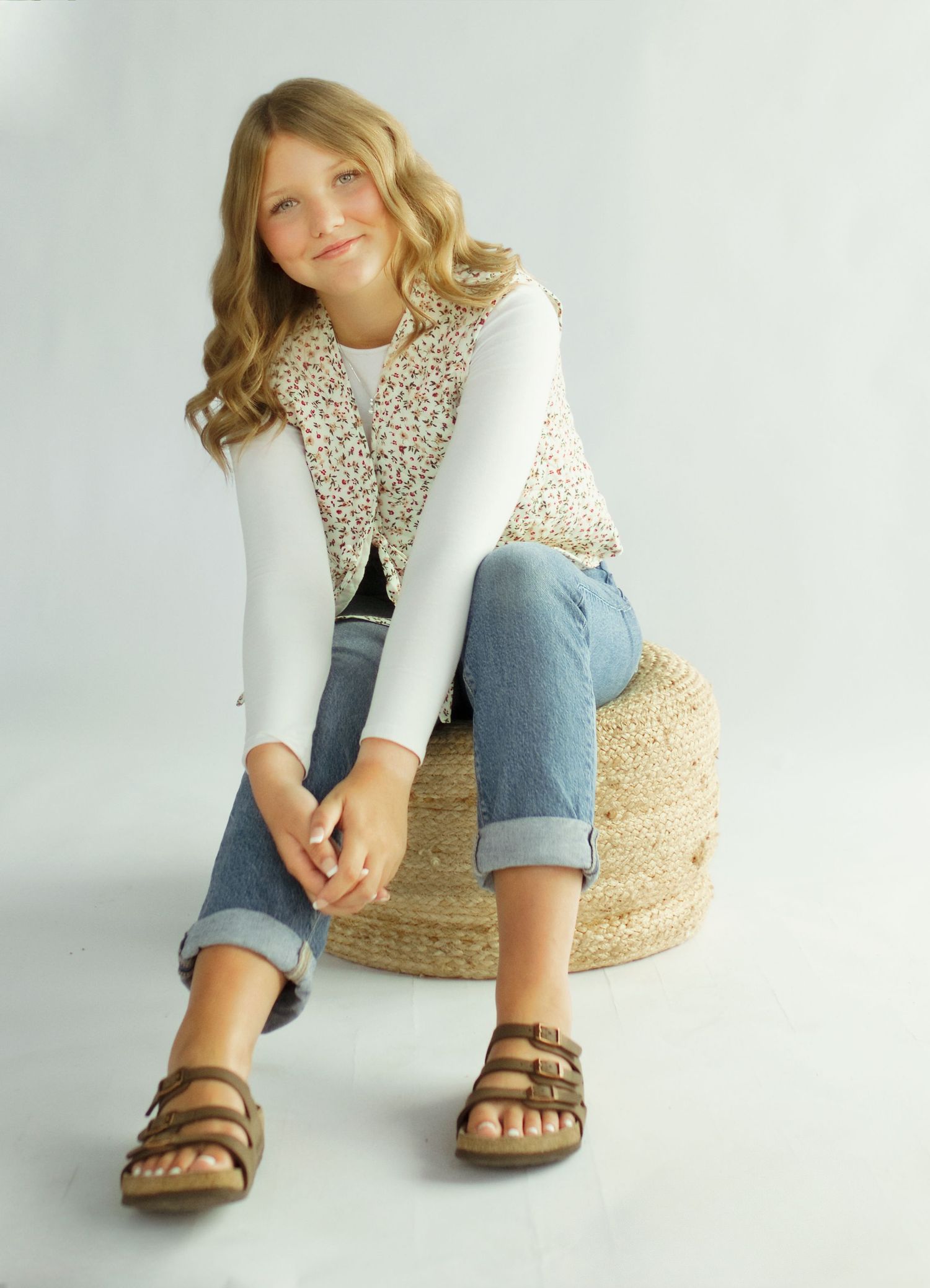 Young girl in floral vest, white top, jeans, and sandals sitting on a woven stool in a studio.