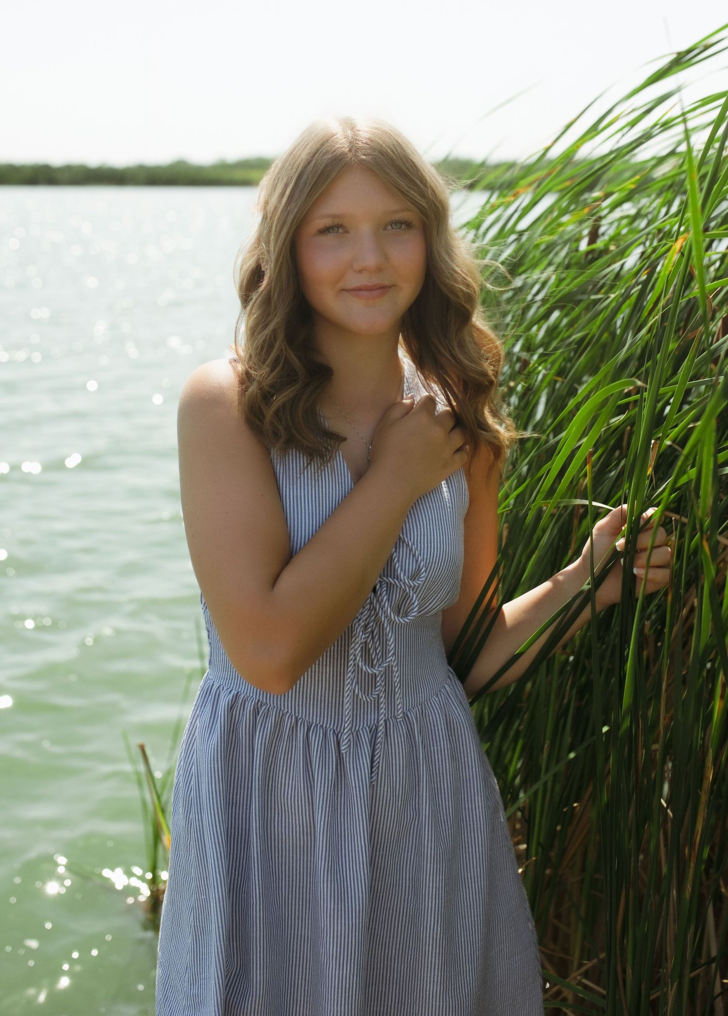 Young girl in blue sundress posing by tall reeds near shimmering water on an overcast day.