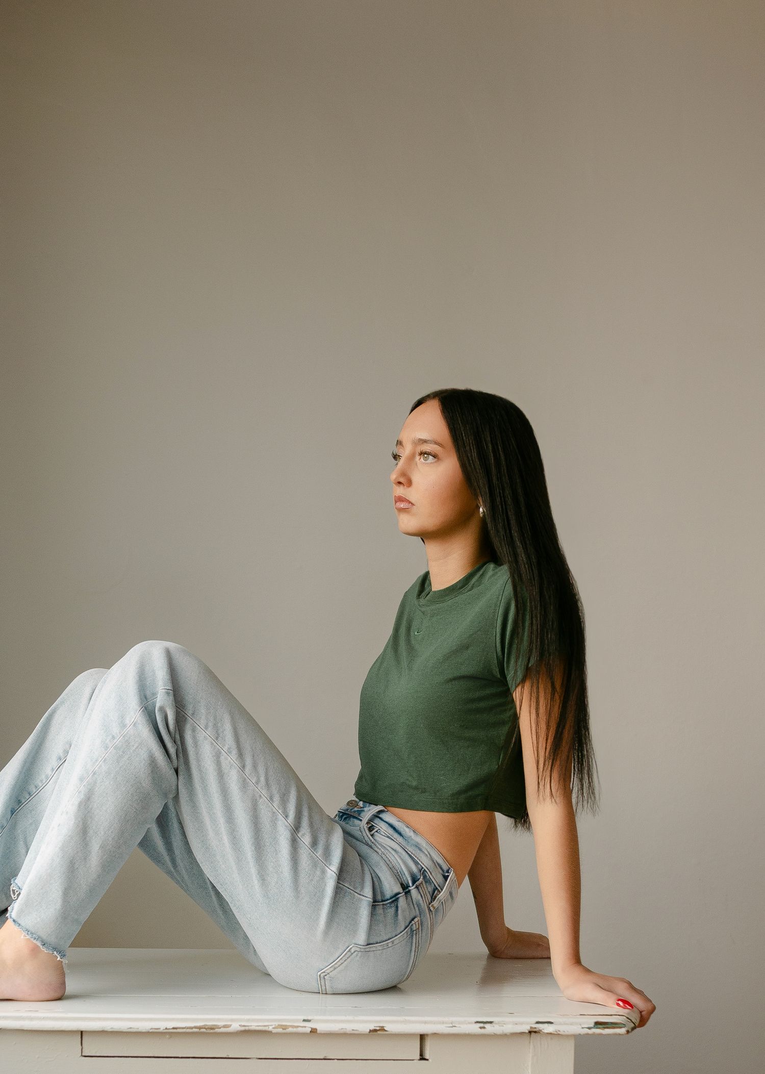 Young woman in green crop top and light wash jeans sitting on white surface against neutral background.