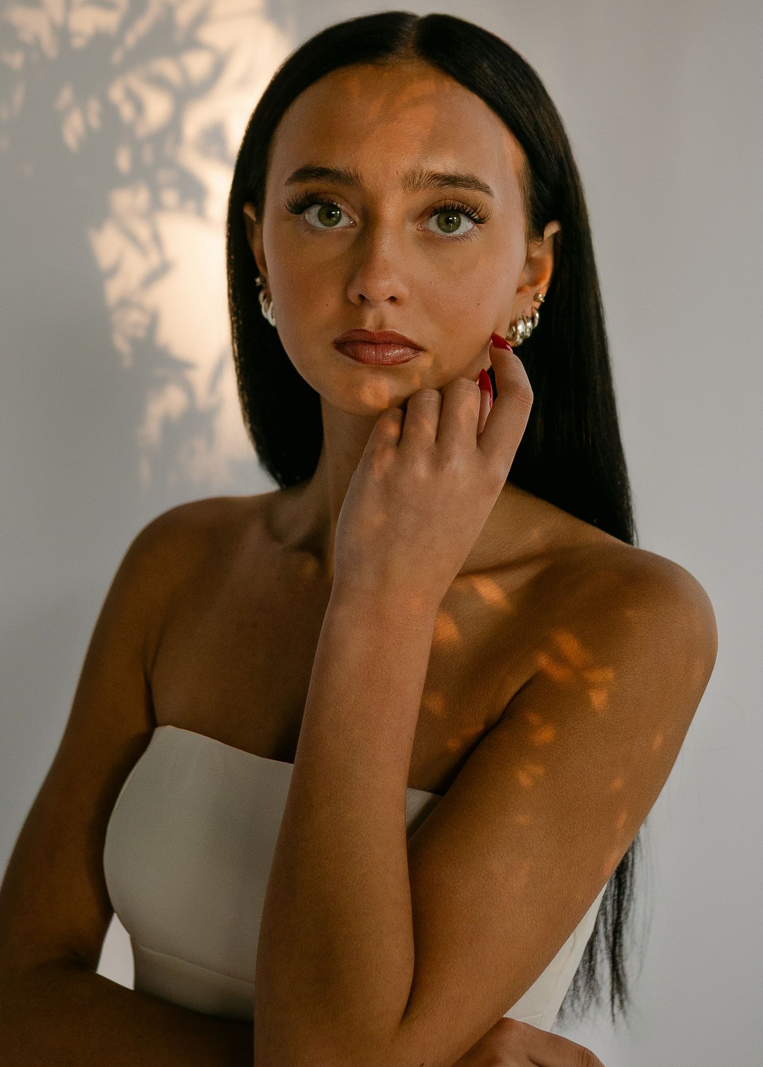 Young woman with long dark hair and green eyes poses gracefully, wearing gold hoop earrings and a white strapless top.