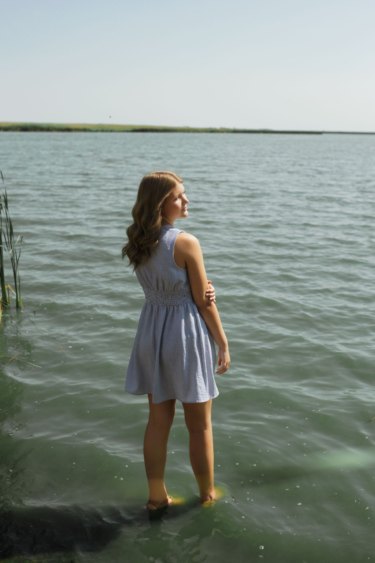 Young woman in blue dress standing in shallow lake water, looking over her shoulder on a clear sunny day.