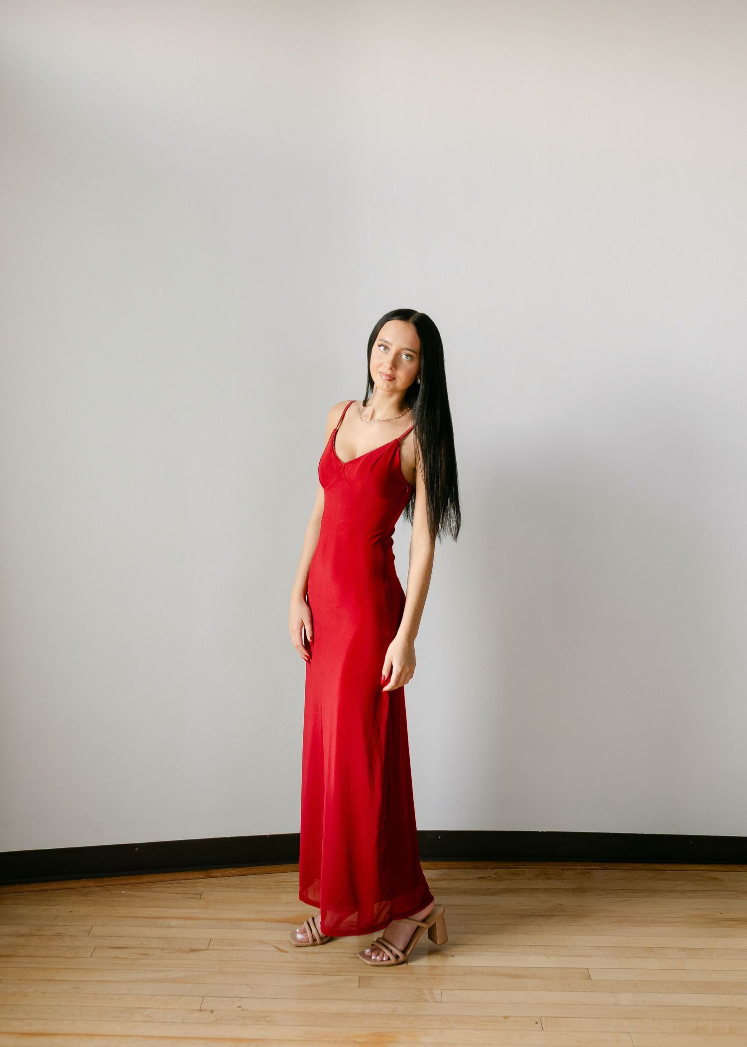 Woman in red spaghetti strap maxi dress and nude heels posing on wood floor against white wall.