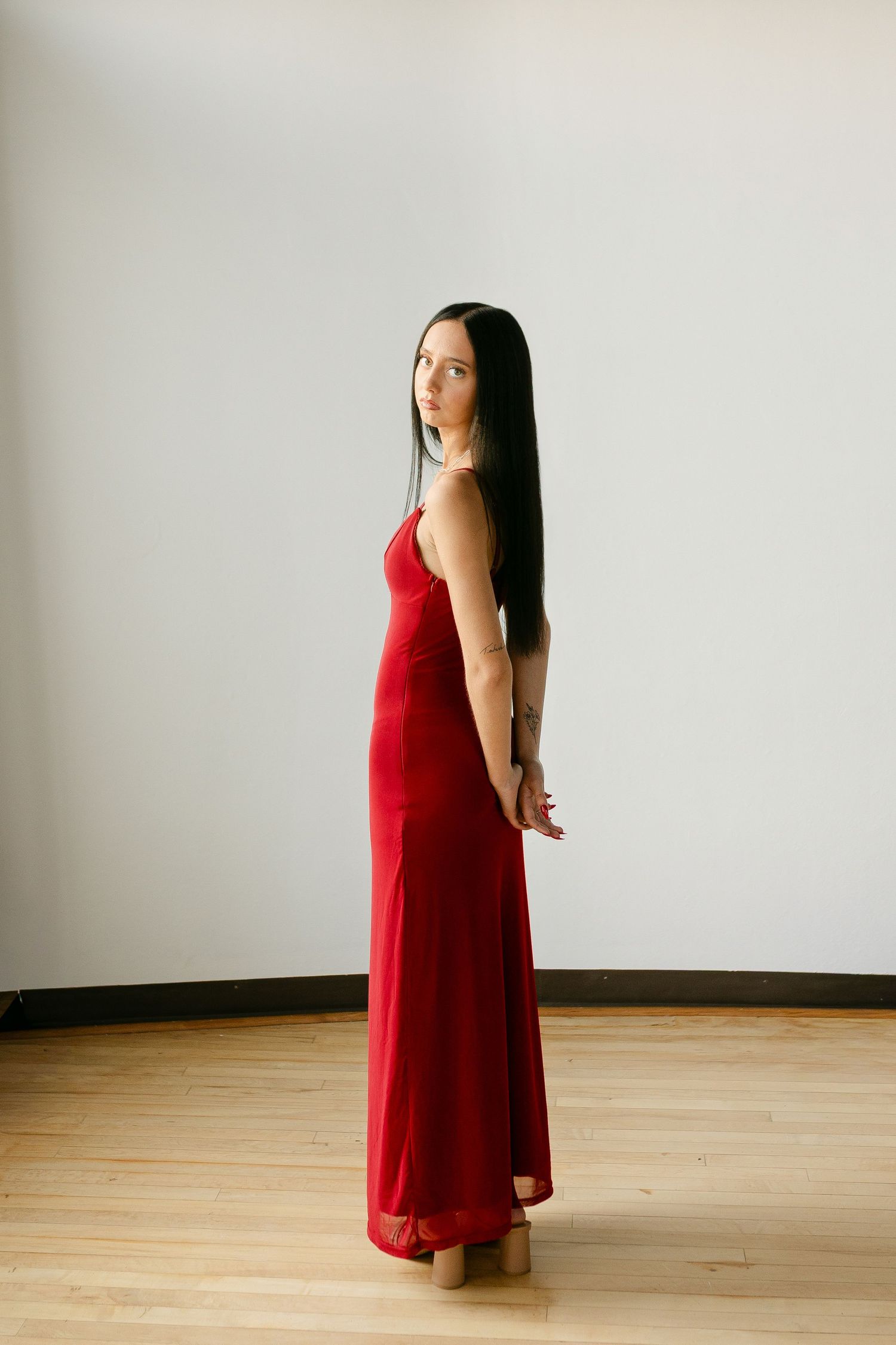 Woman in a long red spaghetti strap dress with long black hair posing in a minimalist studio setting.