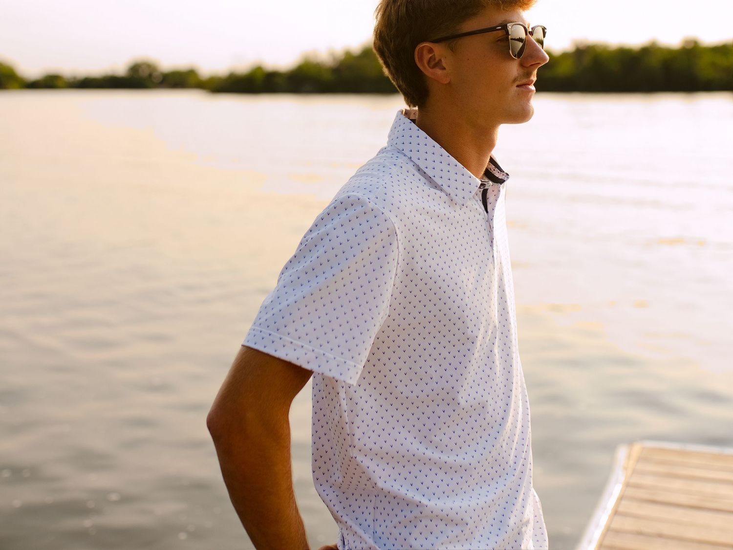 Young man in white patterned shirt and sunglasses standing on a dock by a calm lake at sunset.