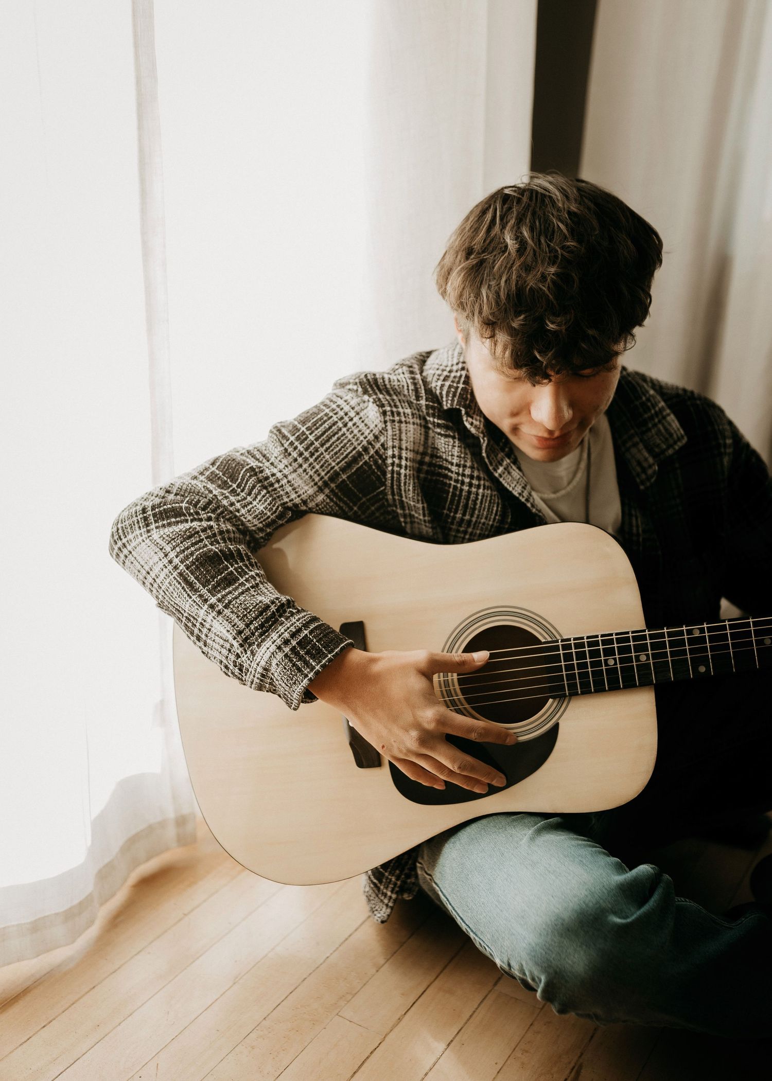 Person playing acoustic guitar while sitting on hardwood floor near white curtains at clark sd rental studio