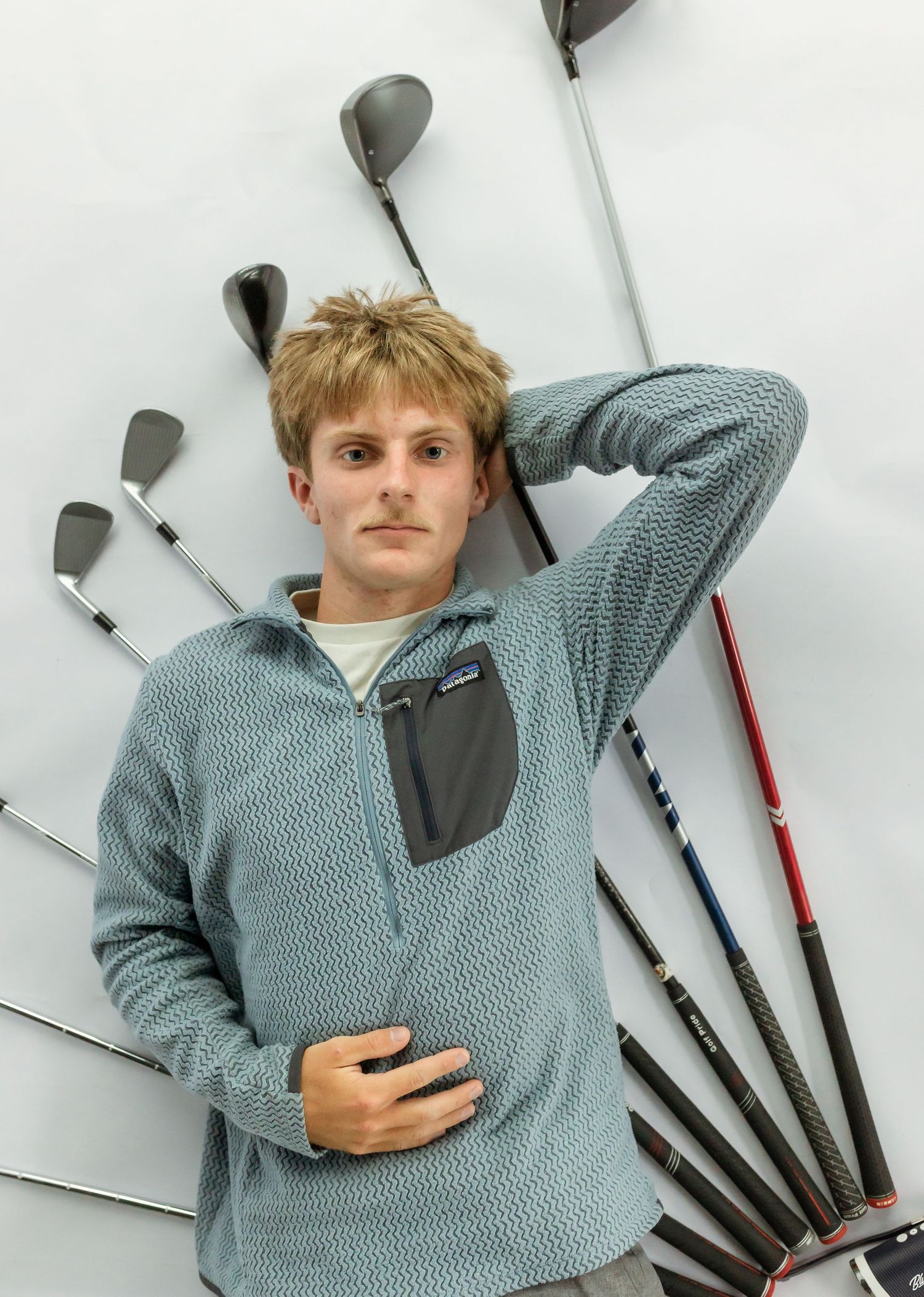 Young man in gray Patagonia fleece pullover posing with multiple golf clubs fanned out against a white wall.