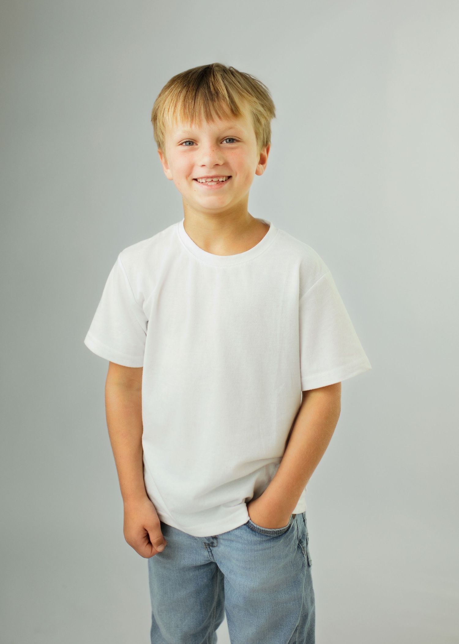 Smiling blonde boy in white t-shirt and jeans posing with hands in pockets against gray background.
