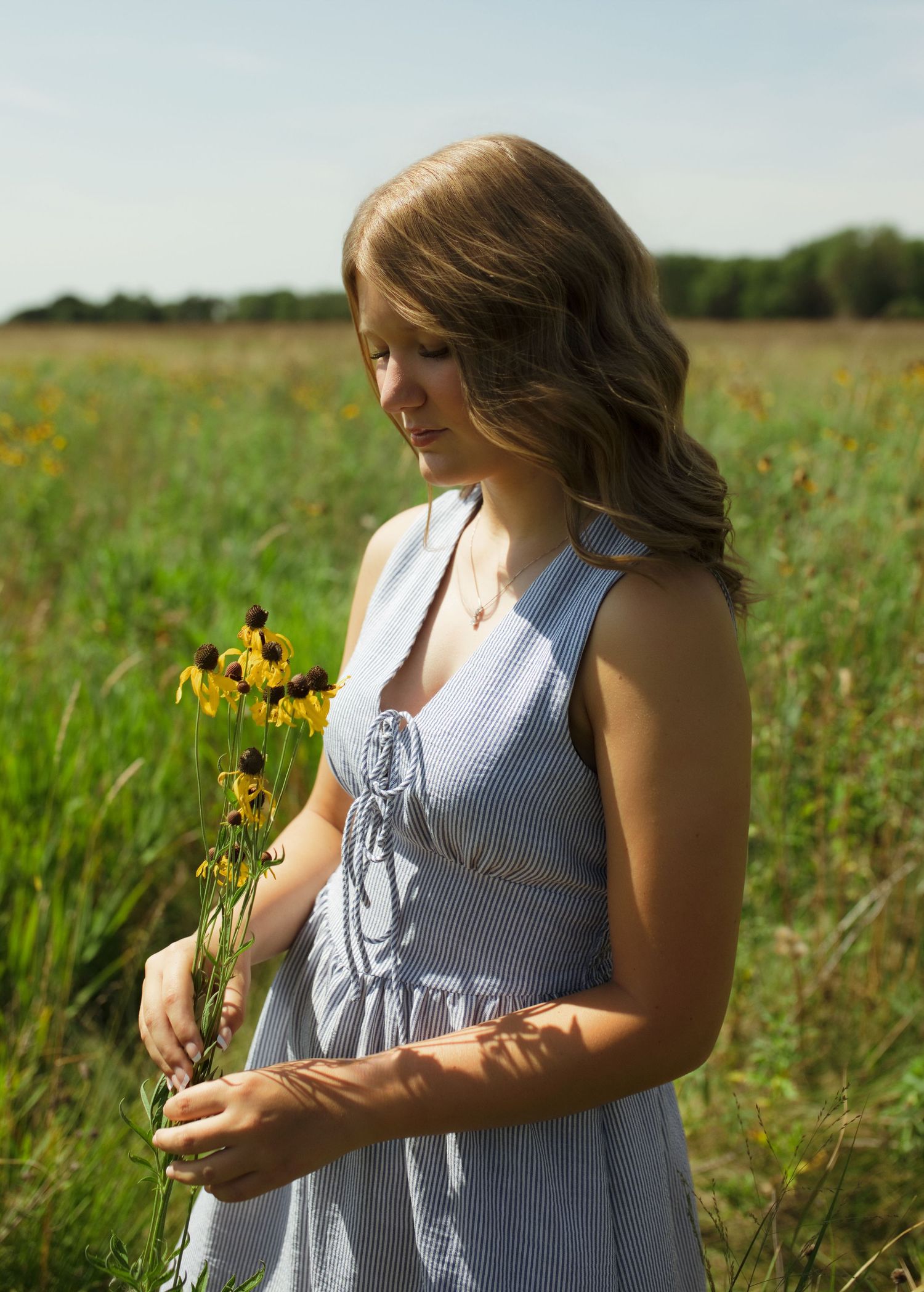 Young woman in blue striped dress holding yellow wildflowers in a sunny green meadow.