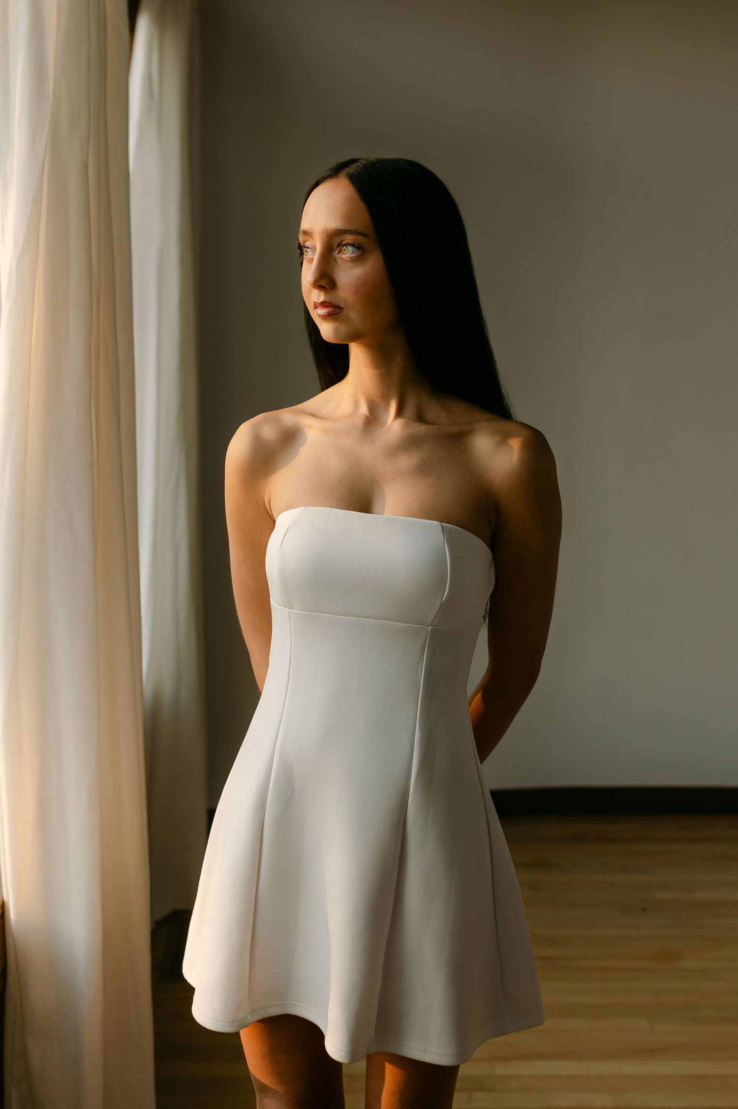 Young woman with long dark hair wearing a white strapless mini dress, standing near a curtained window.