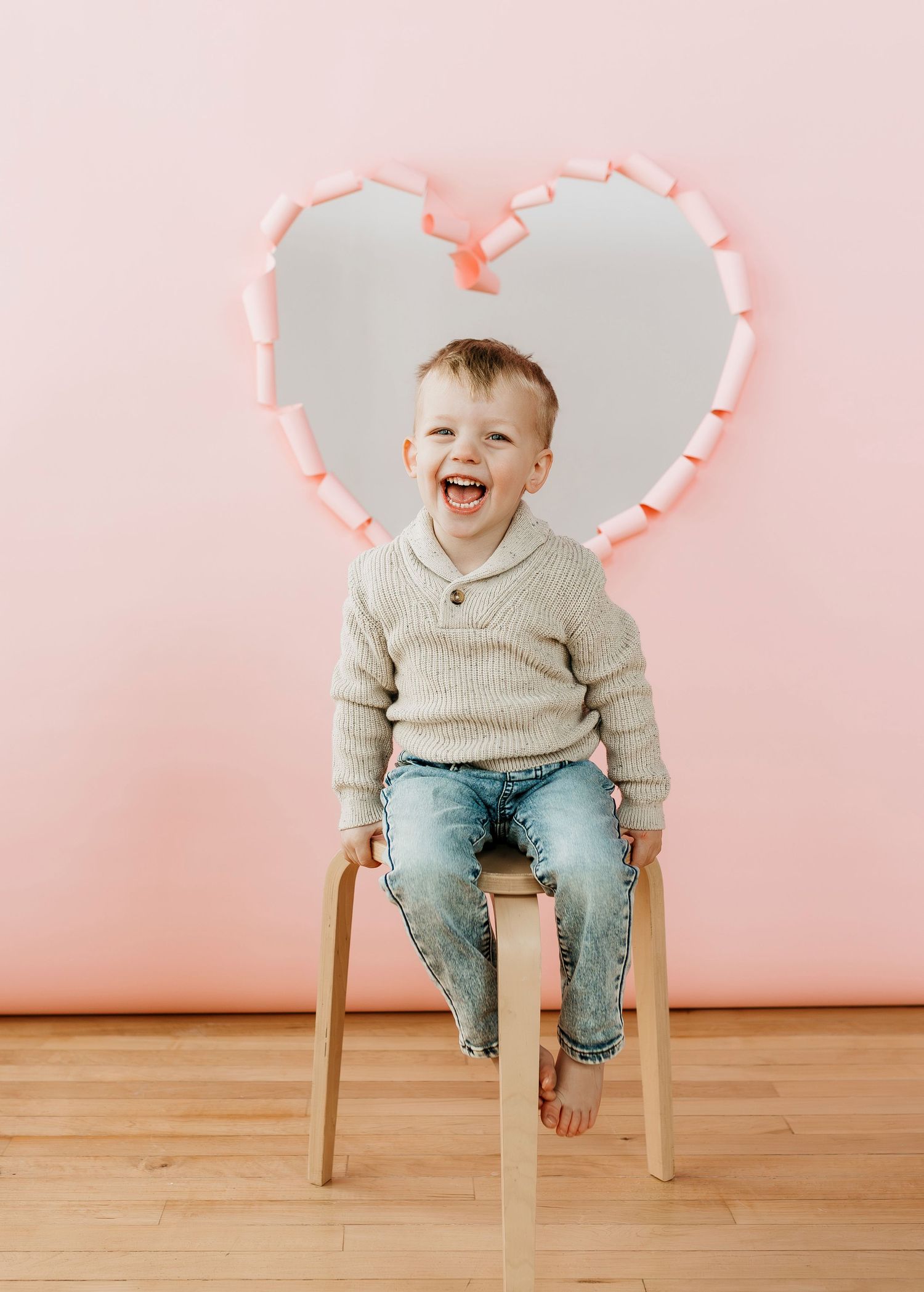 Laughing child in beige sweater sits on wooden chair against pink background with paper heart frame.