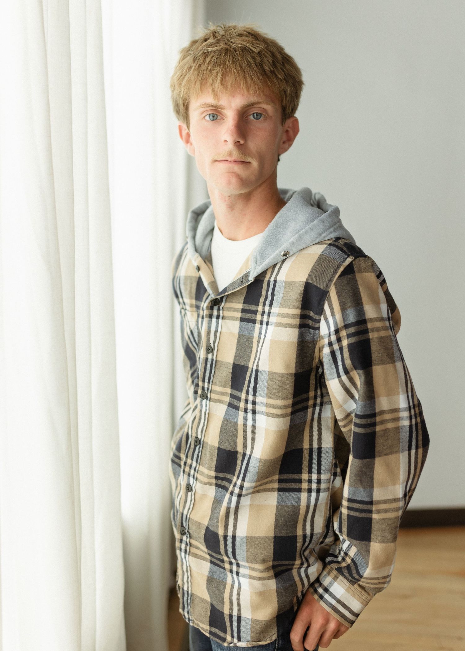 Young man in plaid flannel hoodie shirt standing by a window with natural light.