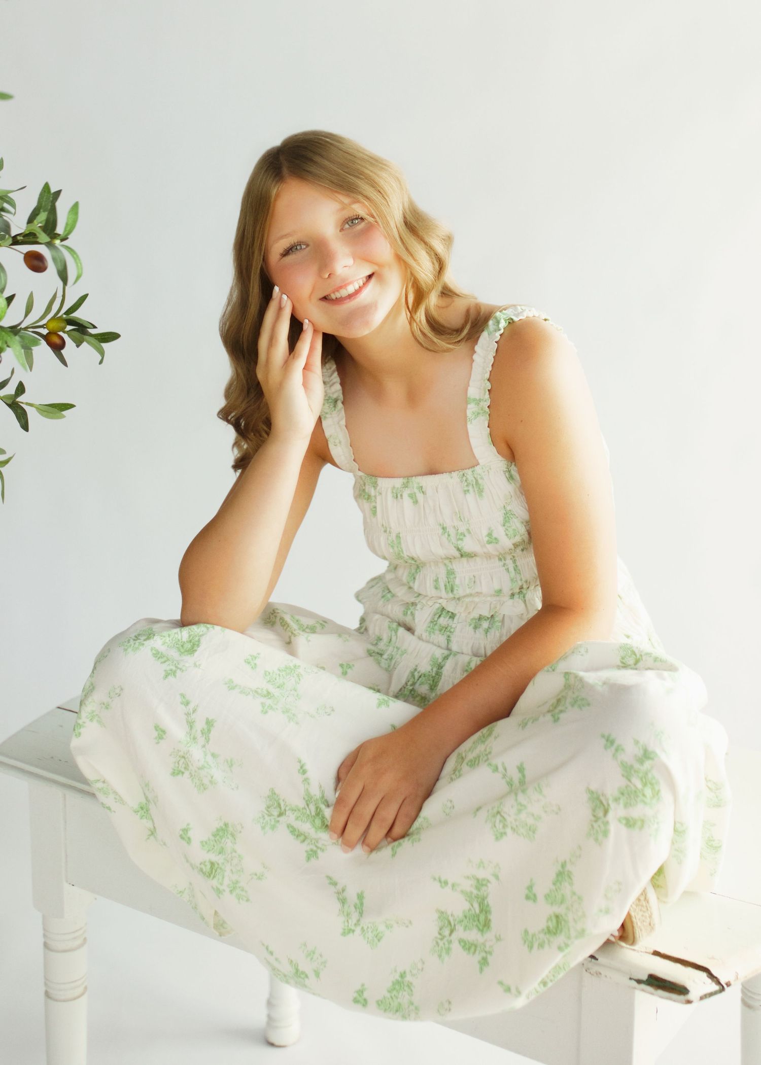 Young girl in a white and green floral sundress sits gracefully on a white bench in a bright studio setting.