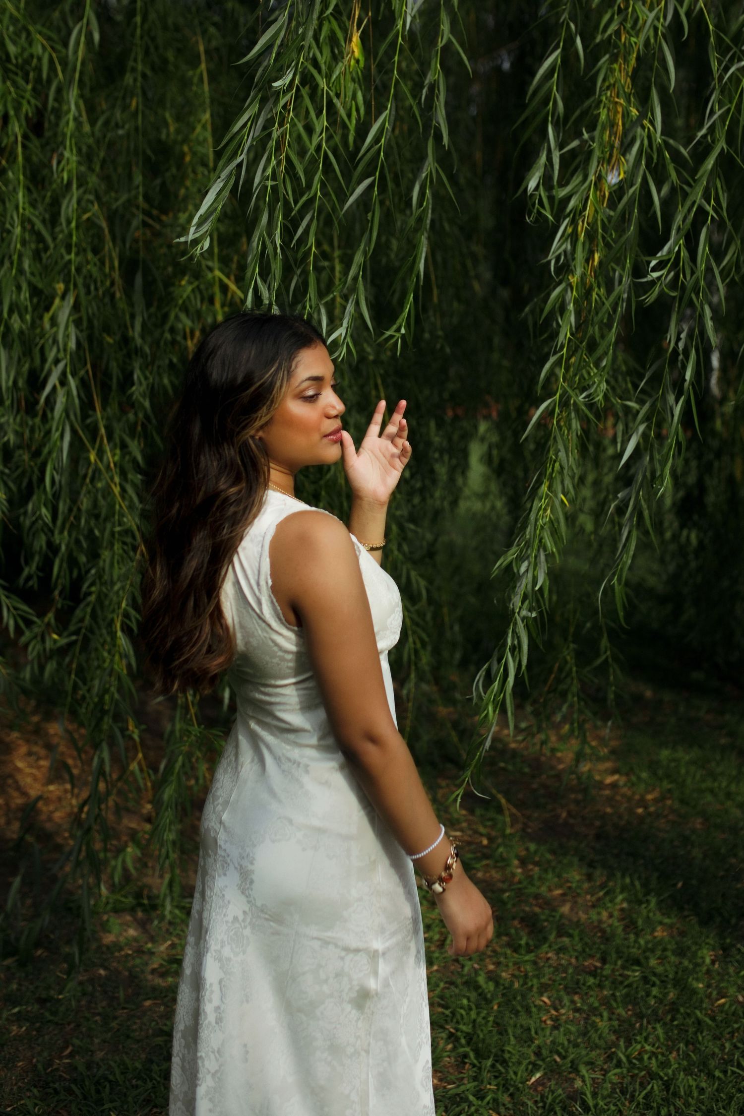 Woman in white dress standing under weeping willow tree, eyes closed, touching branches gracefully.