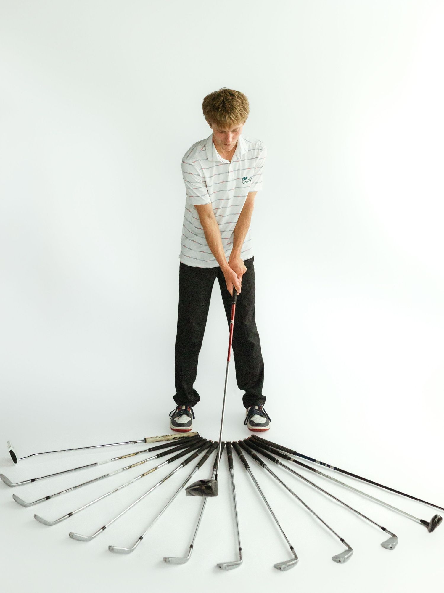 Young golfer holding a club surrounded by a fan of golf irons arranged on the floor in a studio setting.
