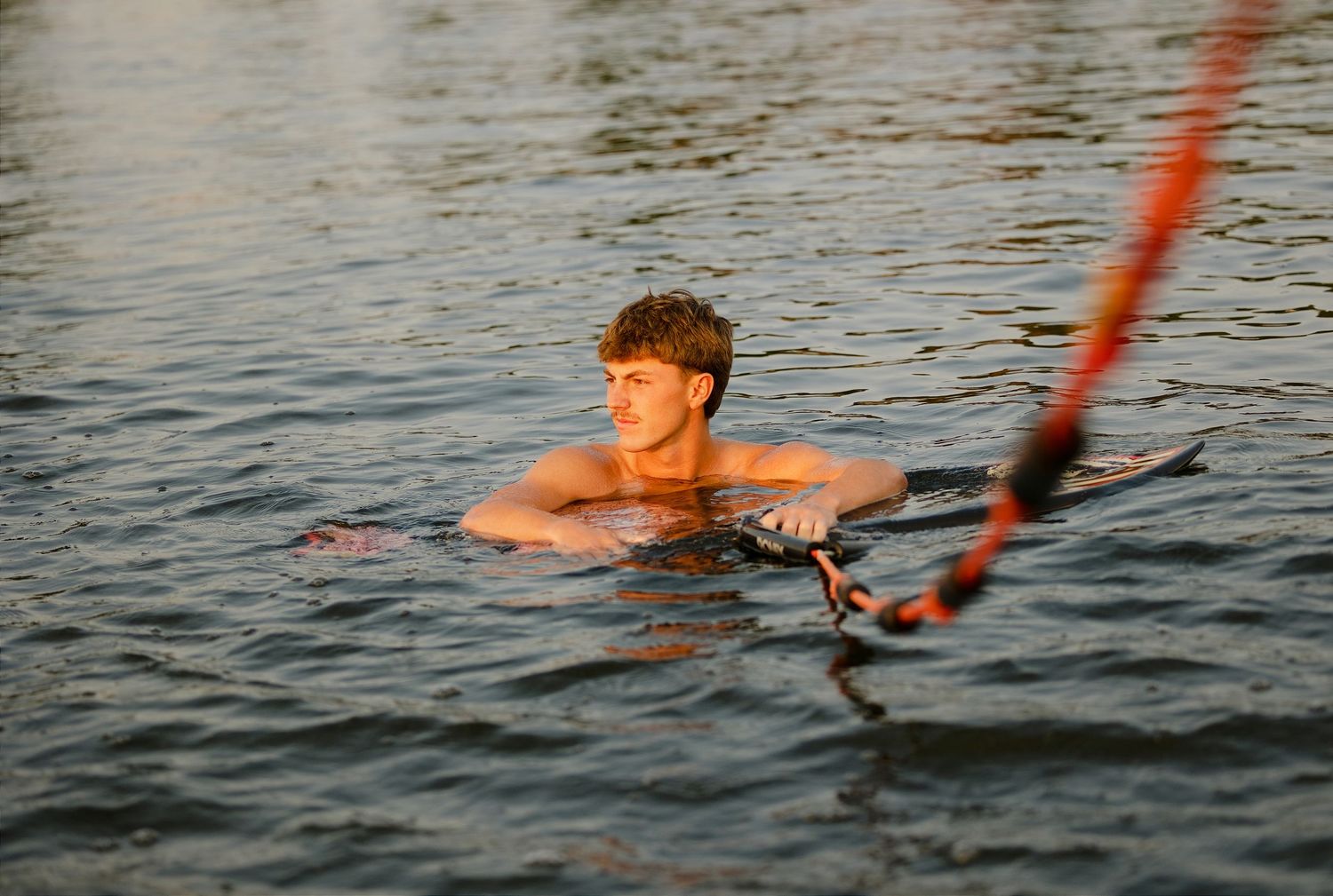 Young man swimming in calm water while holding onto a red rope during golden hour light.