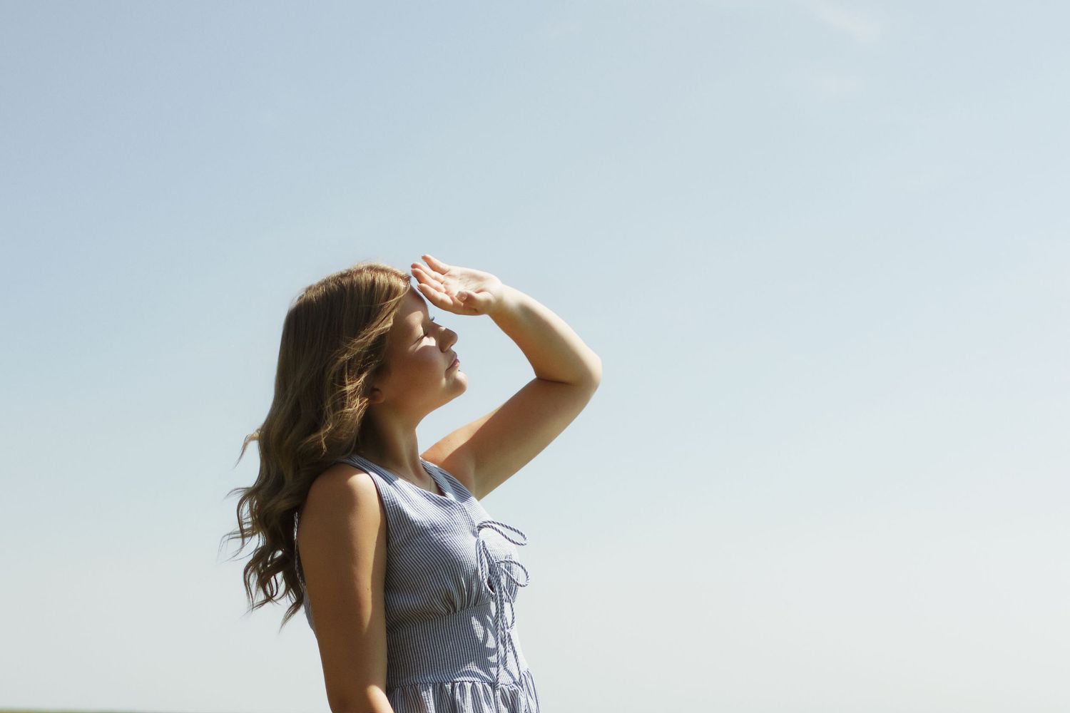Young woman in blue dress shielding eyes from sun against clear blue sky, looking into distance outdoors.