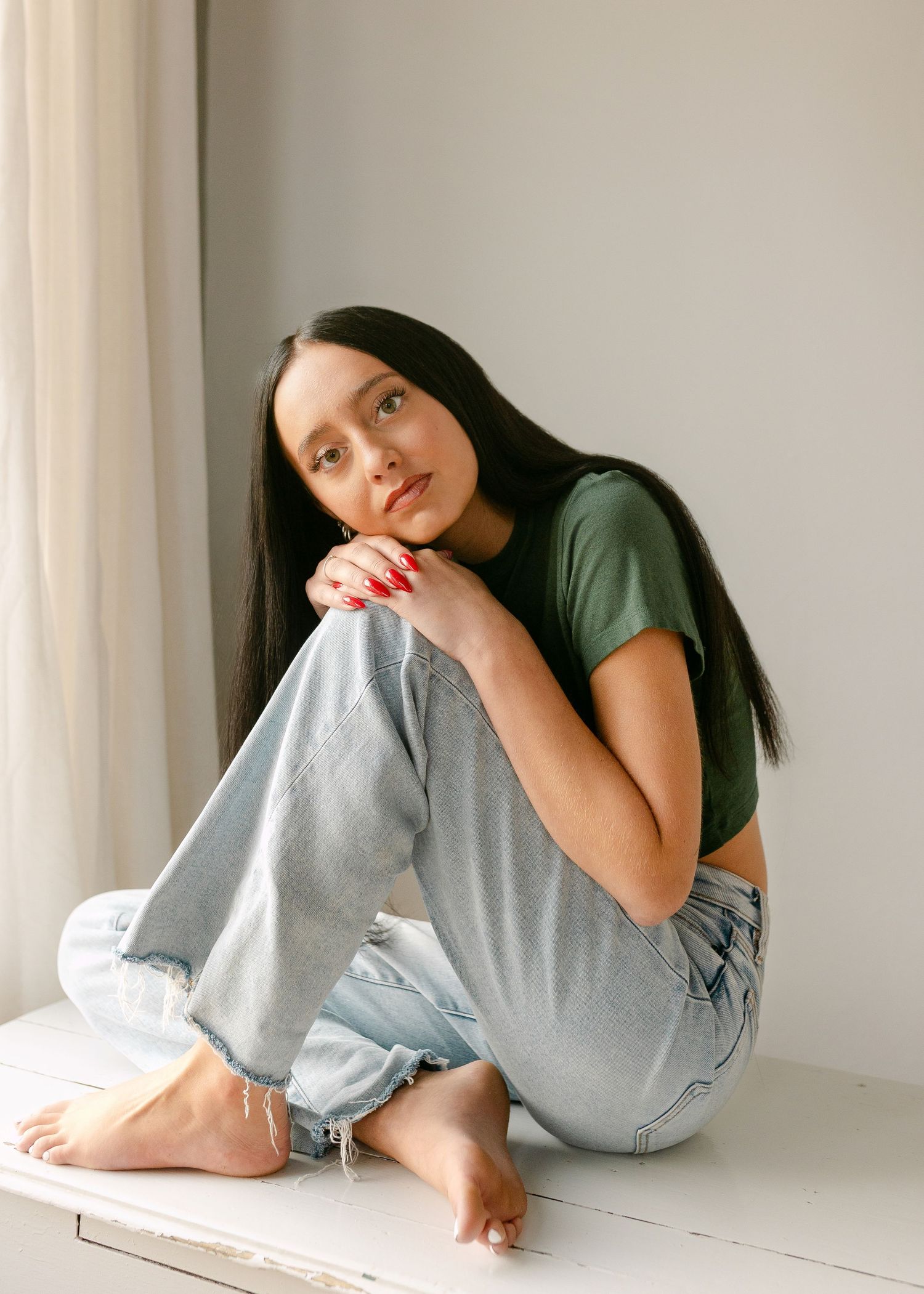 Young woman with long dark hair sits barefoot on white surface wearing green crop top and light wash jeans.