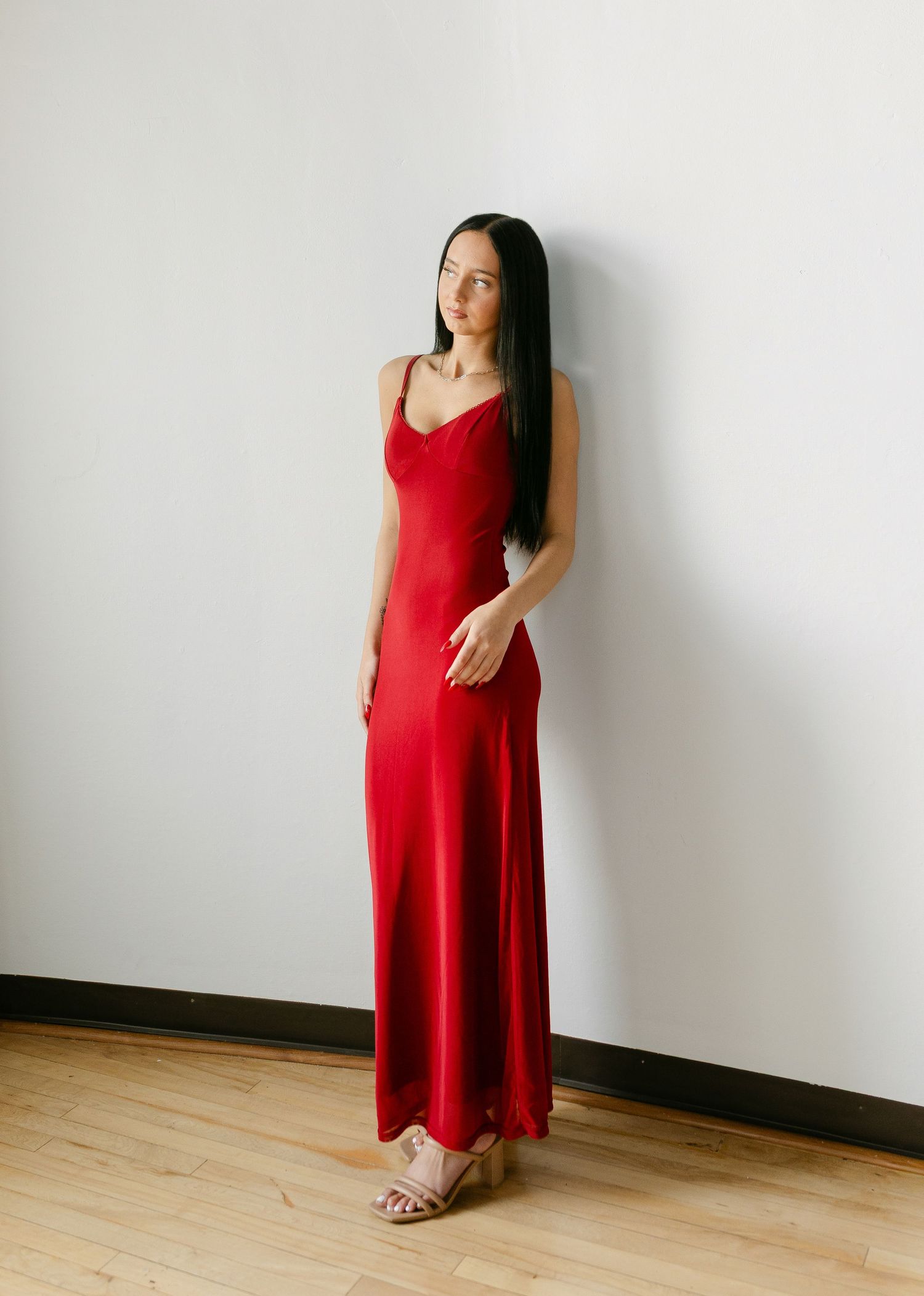 Woman wearing a red satin maxi slip dress with spaghetti straps, standing against a white wall.
