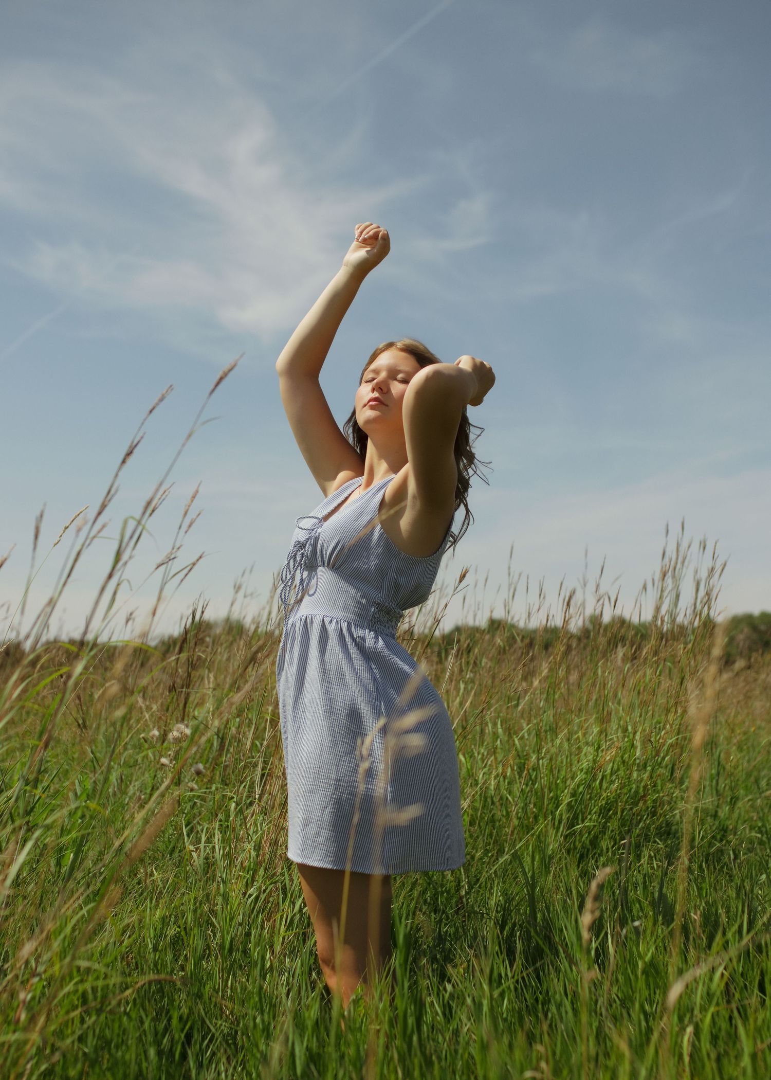 Young woman in blue dress standing in tall green grass field with arms raised under a partly cloudy sky.