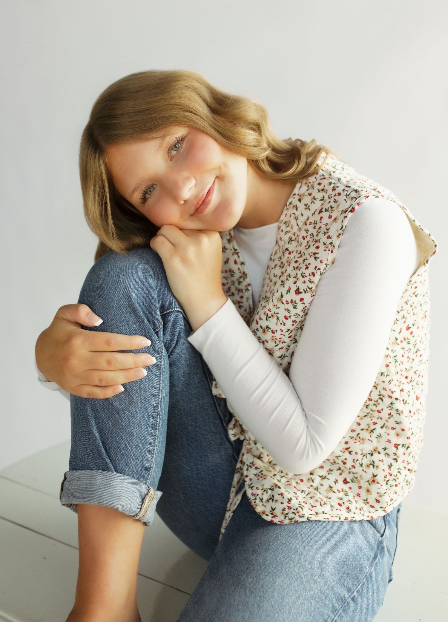 Young girl in floral vest and white long-sleeve top poses casually, resting chin on hand with jeans.