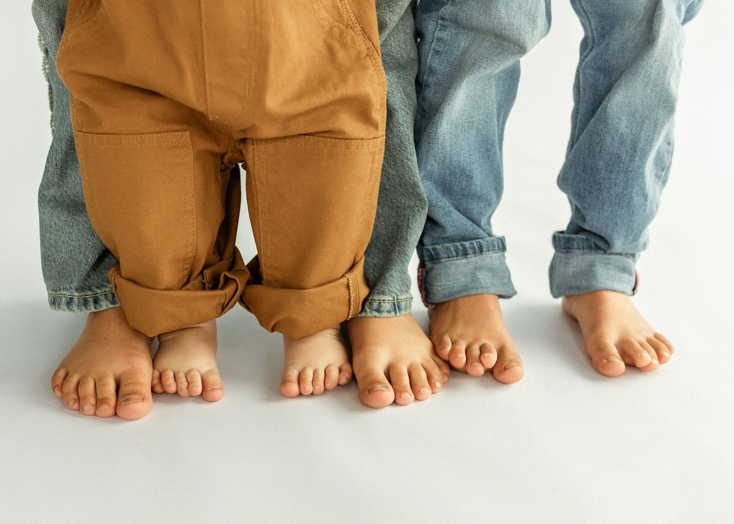 Two barefoot children kneeling and standing on white floor, wearing brown pants and blue jeans.