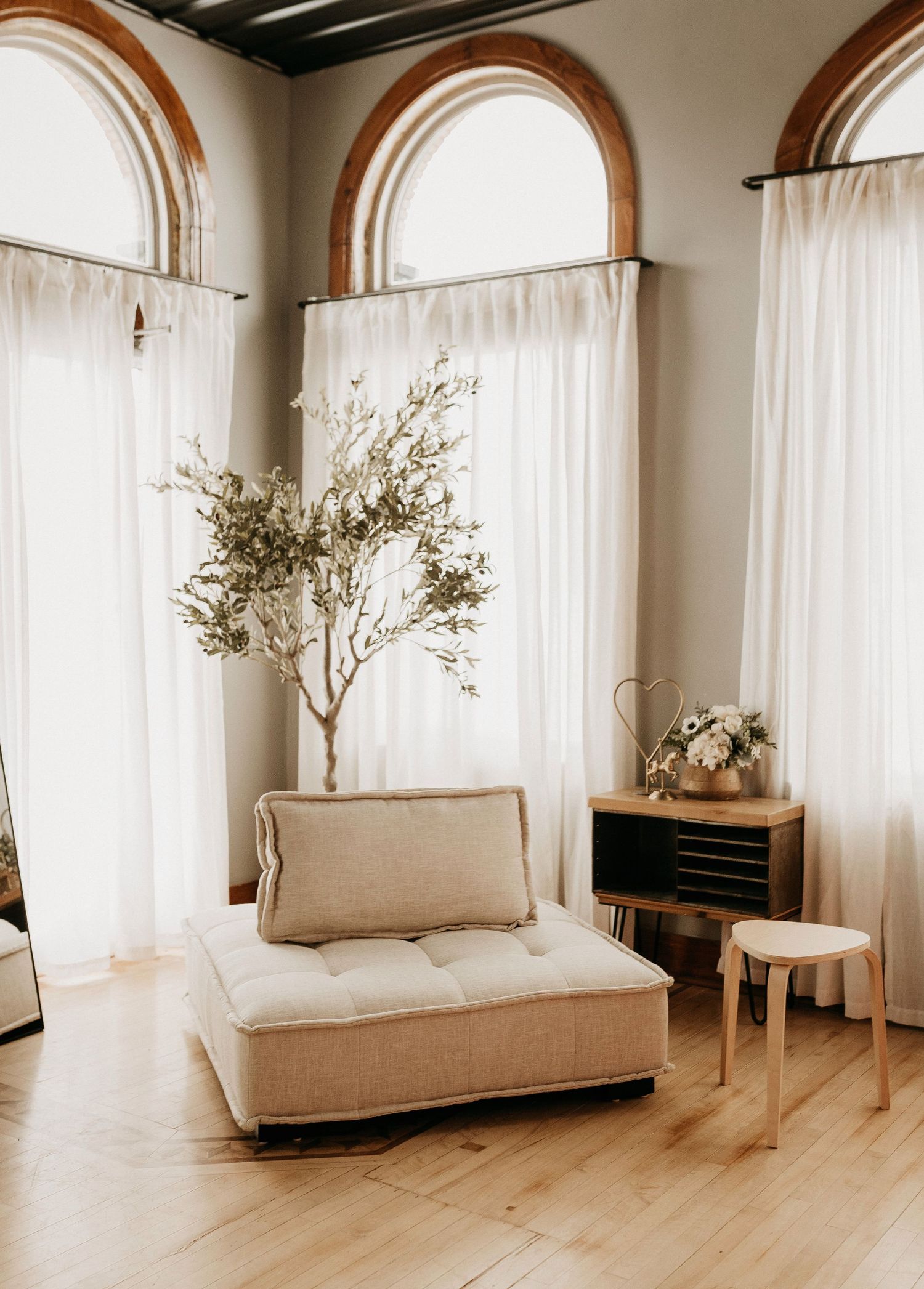 Minimalist living room interior with arched windows, white curtains, and beige furniture on hardwood floors.