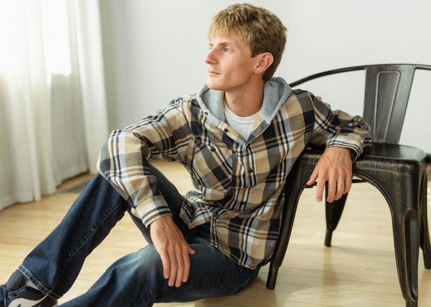Young man in plaid flannel shirt and jeans sitting on floor leaning against metal chair, looking away thoughtfully.