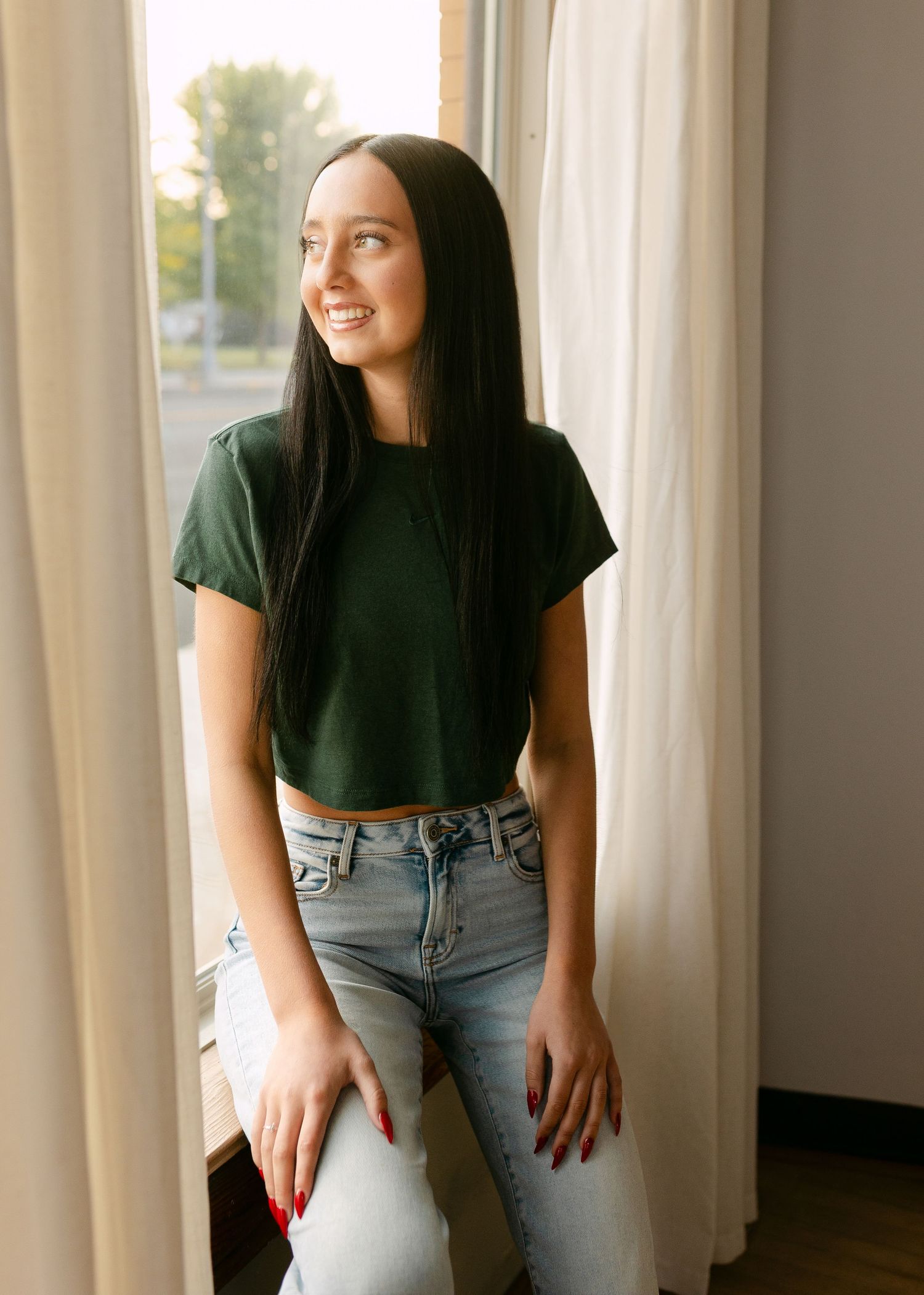 Young woman with long dark hair in green crop top and jeans smiling by a sunlit window with curtains.