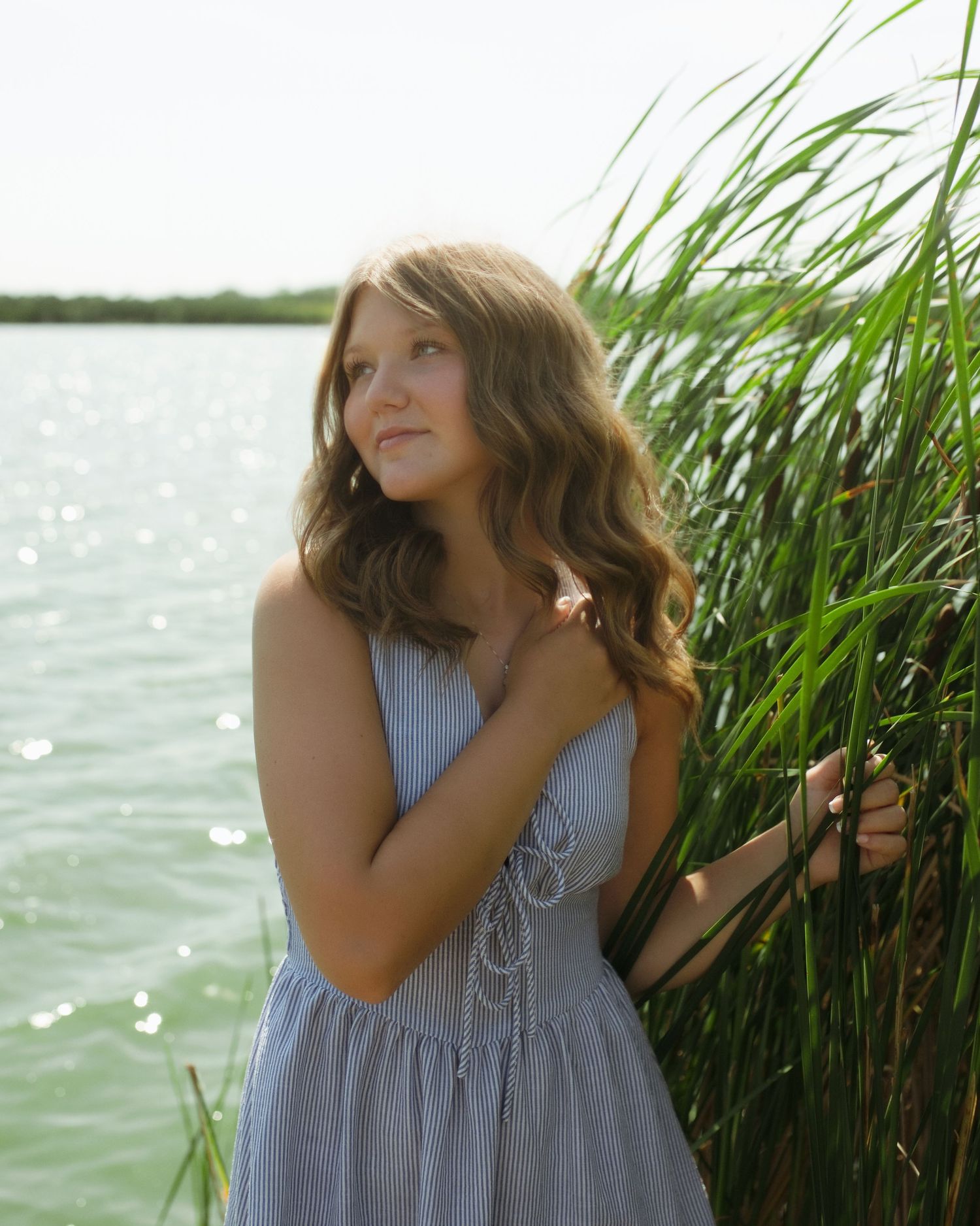 Young woman in blue dress standing by tall green reeds near a shimmering lake, gazing upward dreamily.
