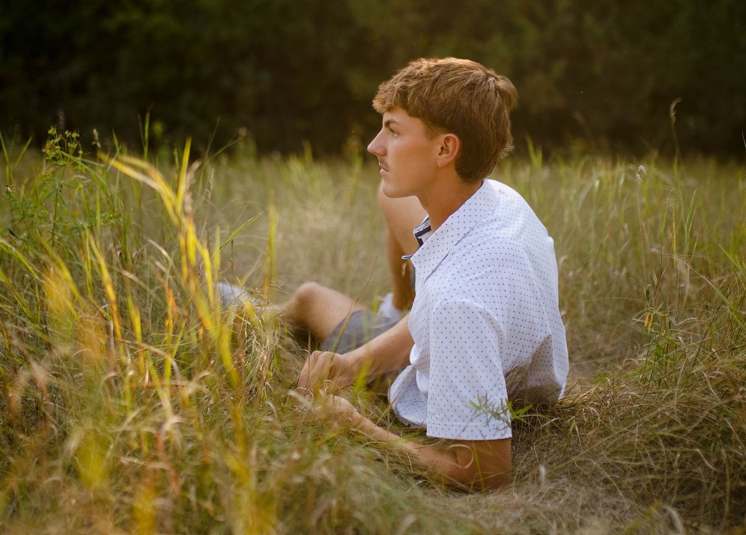 Young man in white shirt sitting in tall golden grass field during golden hour sunset photography.
