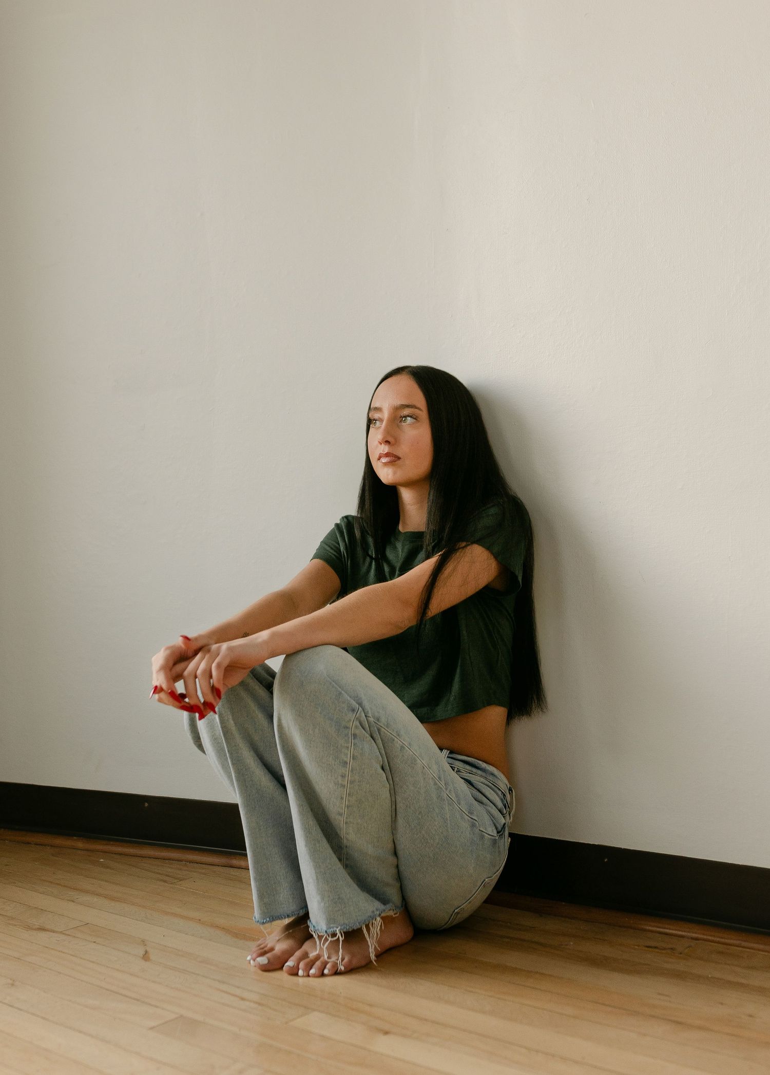 Young woman in green crop top and light jeans sitting against white wall on hardwood floor.