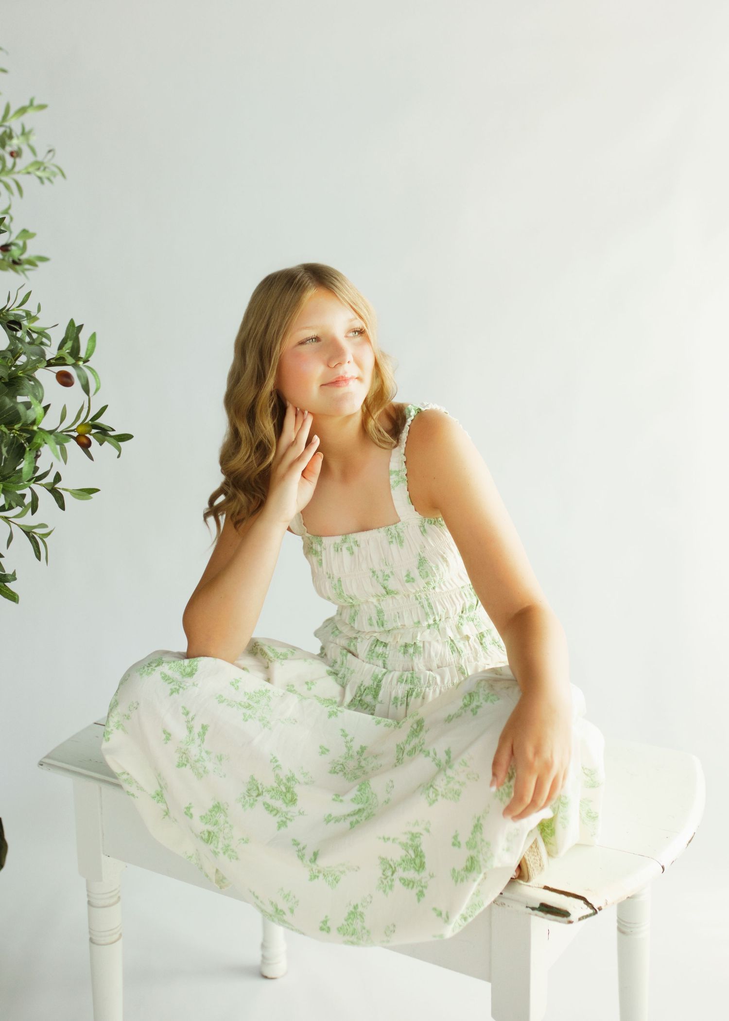 Young girl in white and green floral sundress seated on white bench in bright studio setting.