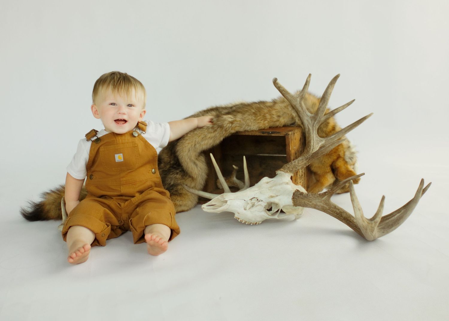 Baby in Carhartt overalls sitting beside deer antlers and animal skull on white background.