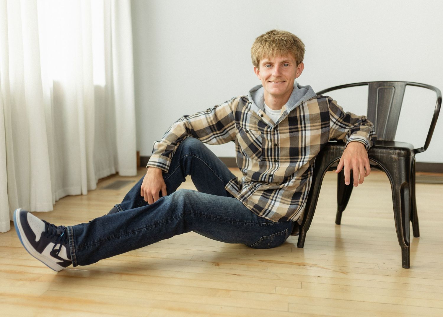 A young man in a plaid shirt and jeans sits casually on a hardwood floor leaning against a metal chair.