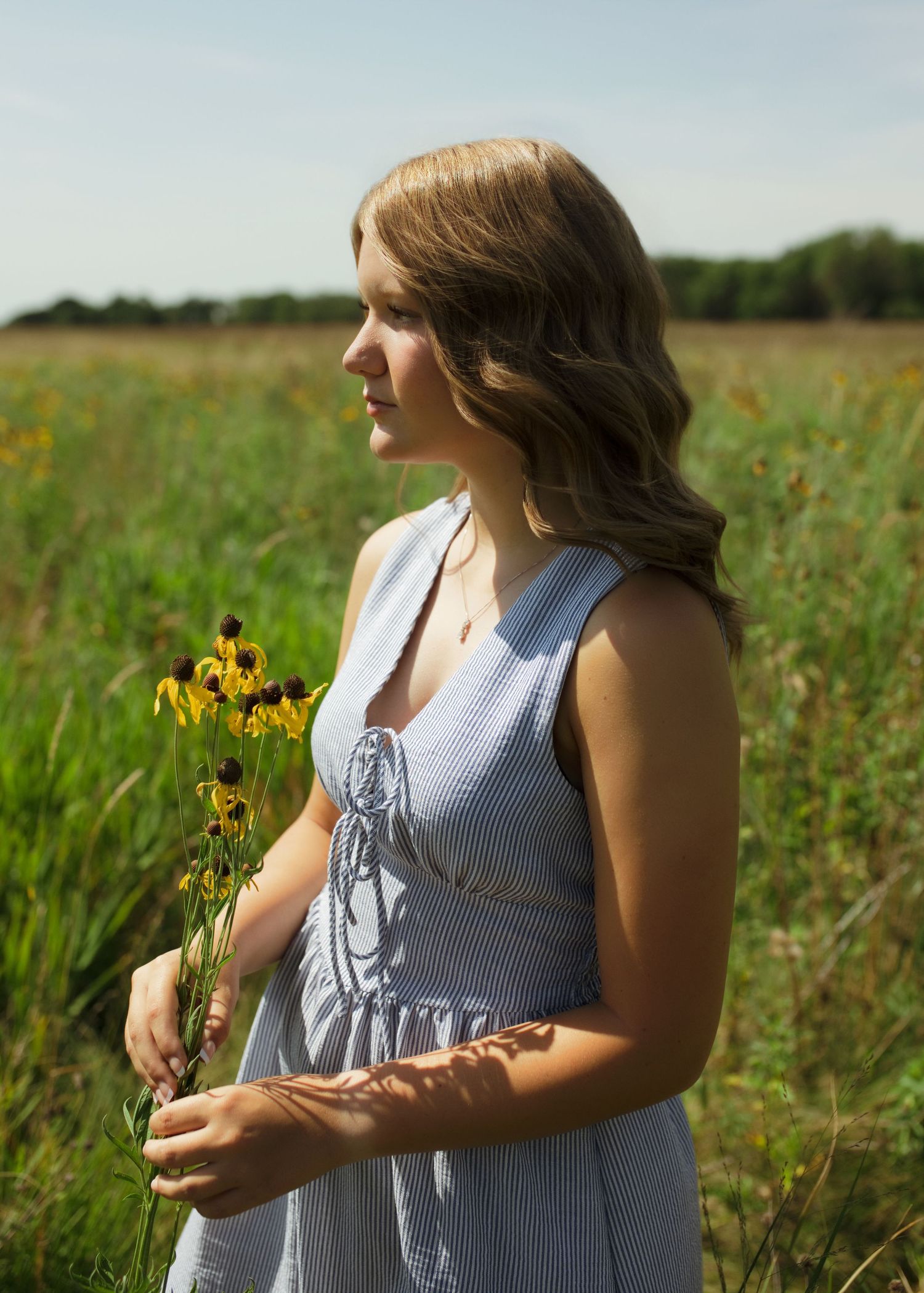 Young woman in blue dress holding yellow wildflowers in a sunny green meadow field.