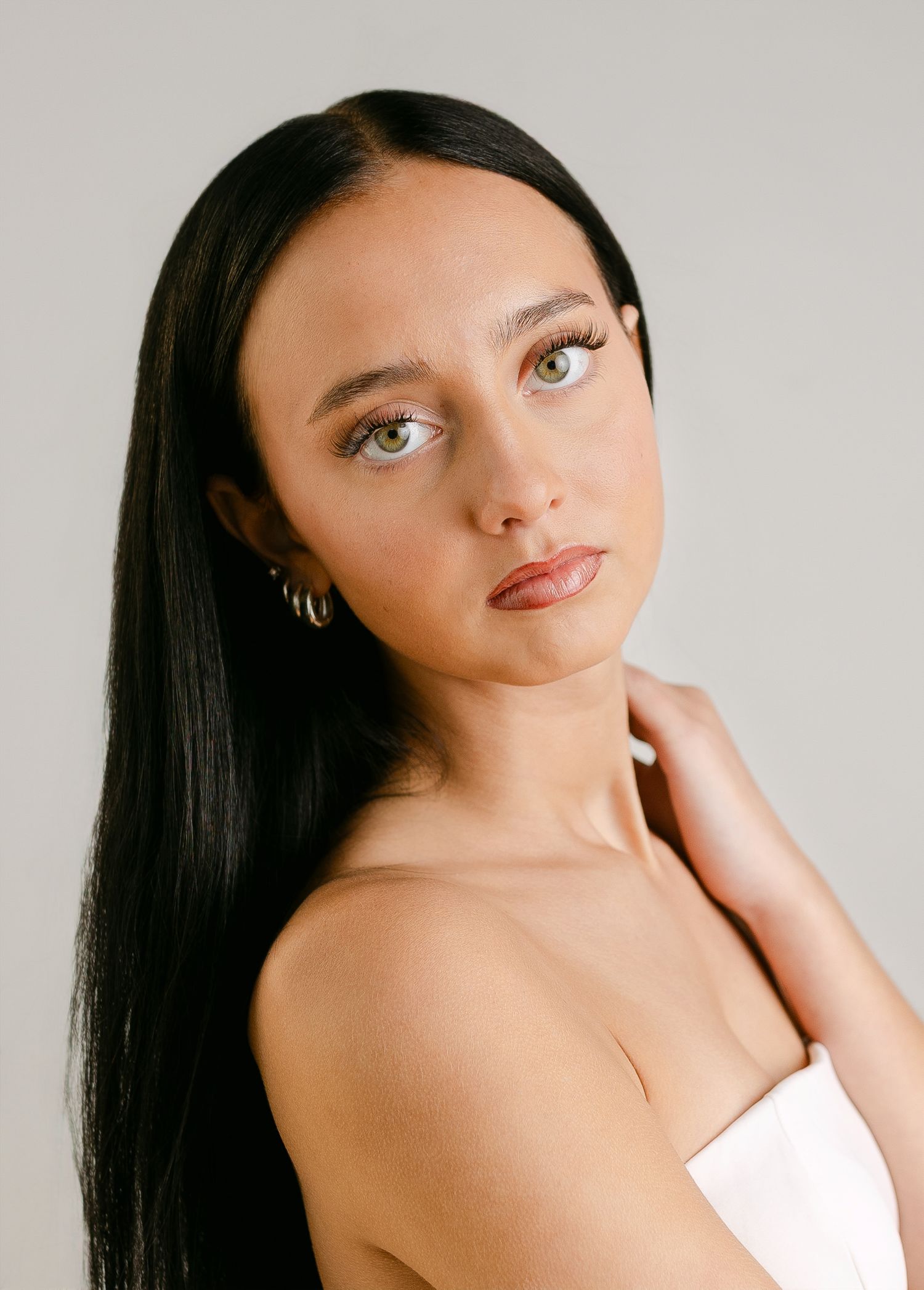 Young woman with long black hair and green eyes wearing gold hoop earrings and a white top poses gracefully.