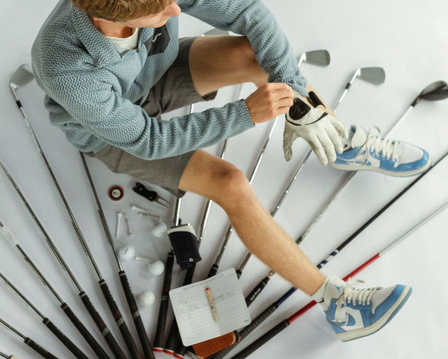 Overhead view of a golfer sitting among golf clubs, wearing blue sneakers and a glove on a white background.