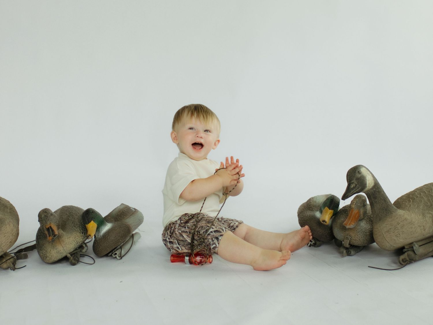 Happy baby clapping hands surrounded by decorative duck decoys on white background.