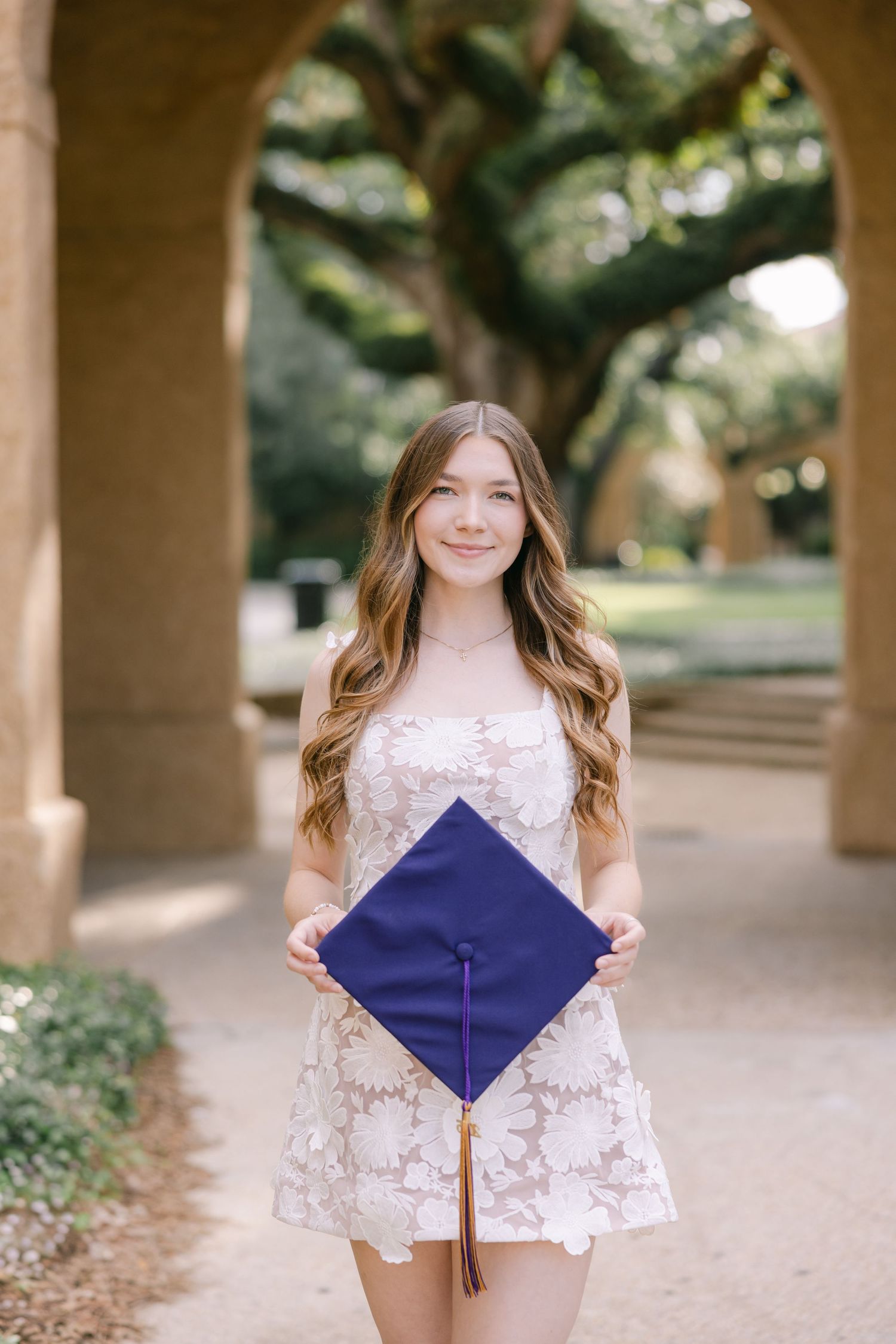 A young college graduate smiles softly at the camera, standing in front of stone arches posing with her graduation cap.
