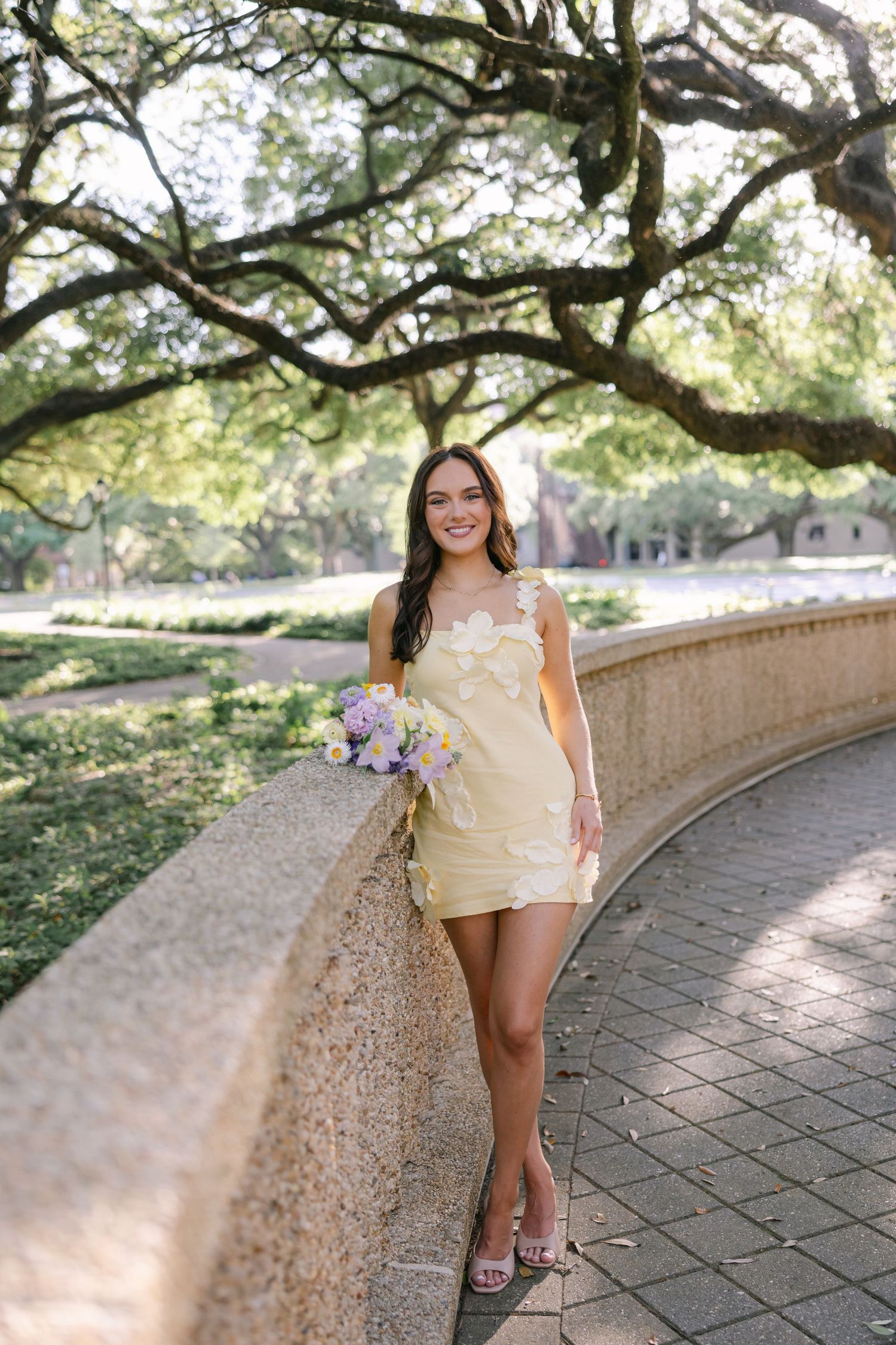 A woman in a floral yellow dress rests against a stone railing. Beautiful oak trees, delicate bouquet LSU GRAD PHOTOGRAPHER