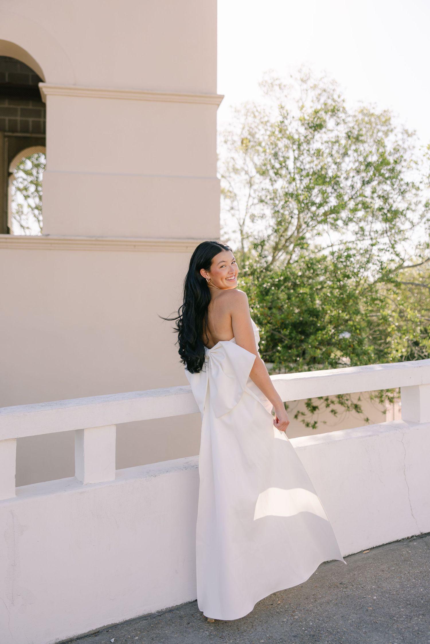 A young woman looks back at the camera, holding a draping bow that cascades down her dress and onto the ground. She stands in front of stone