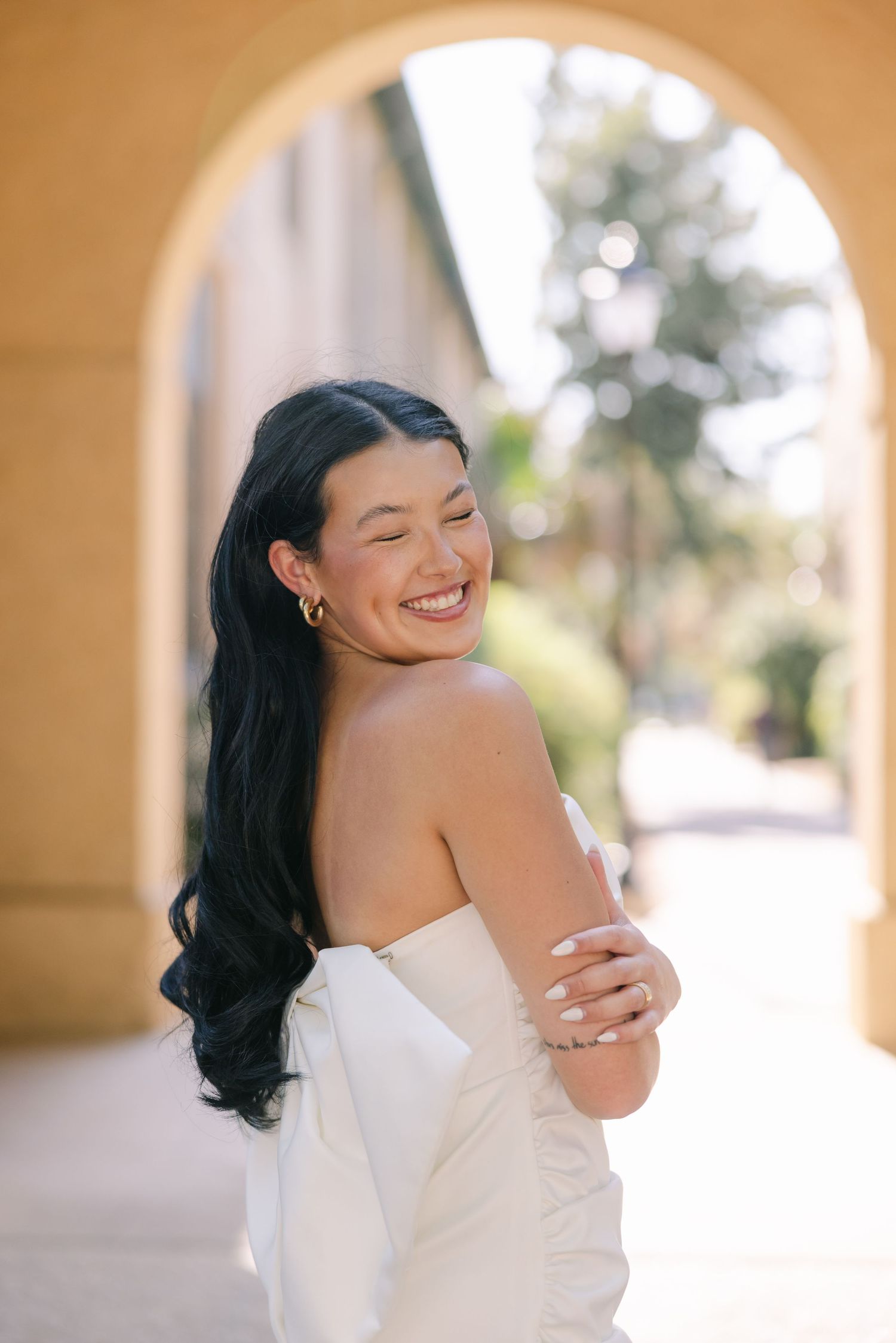 A young woman in front of architectural stone arches smiles and closes her eyes the the camera while caressing her arms