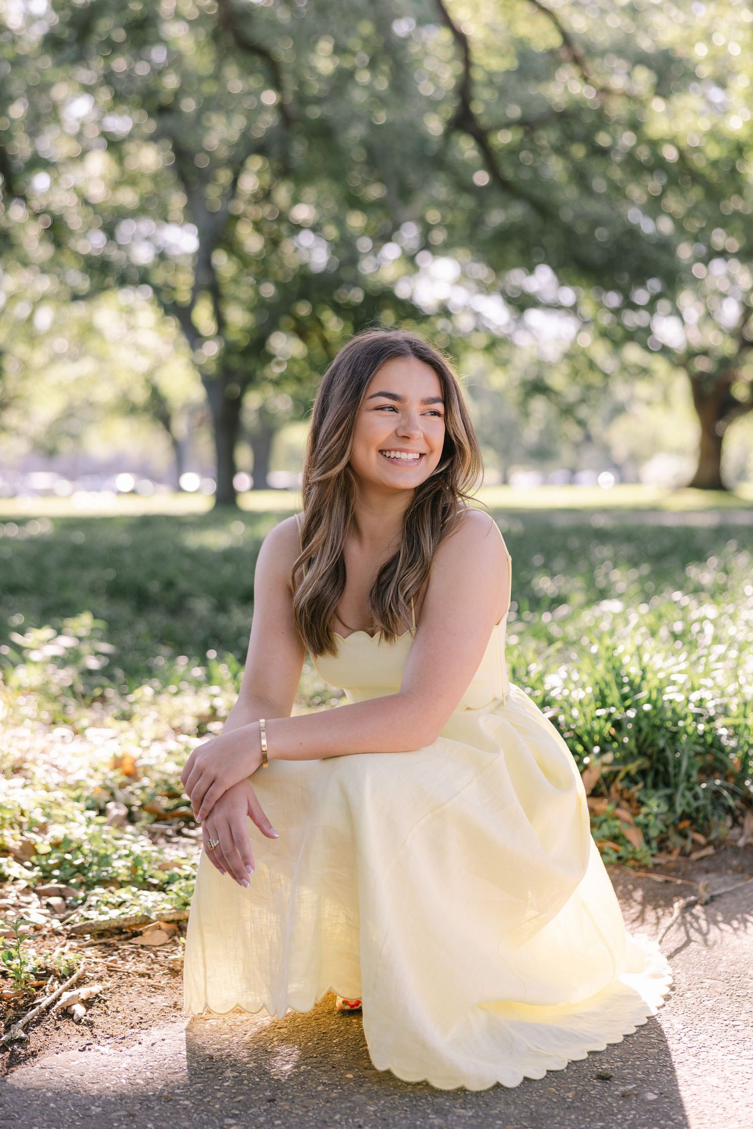 A young woman squats down and leans on her knees and smiles off into the distance in front of historic oaks