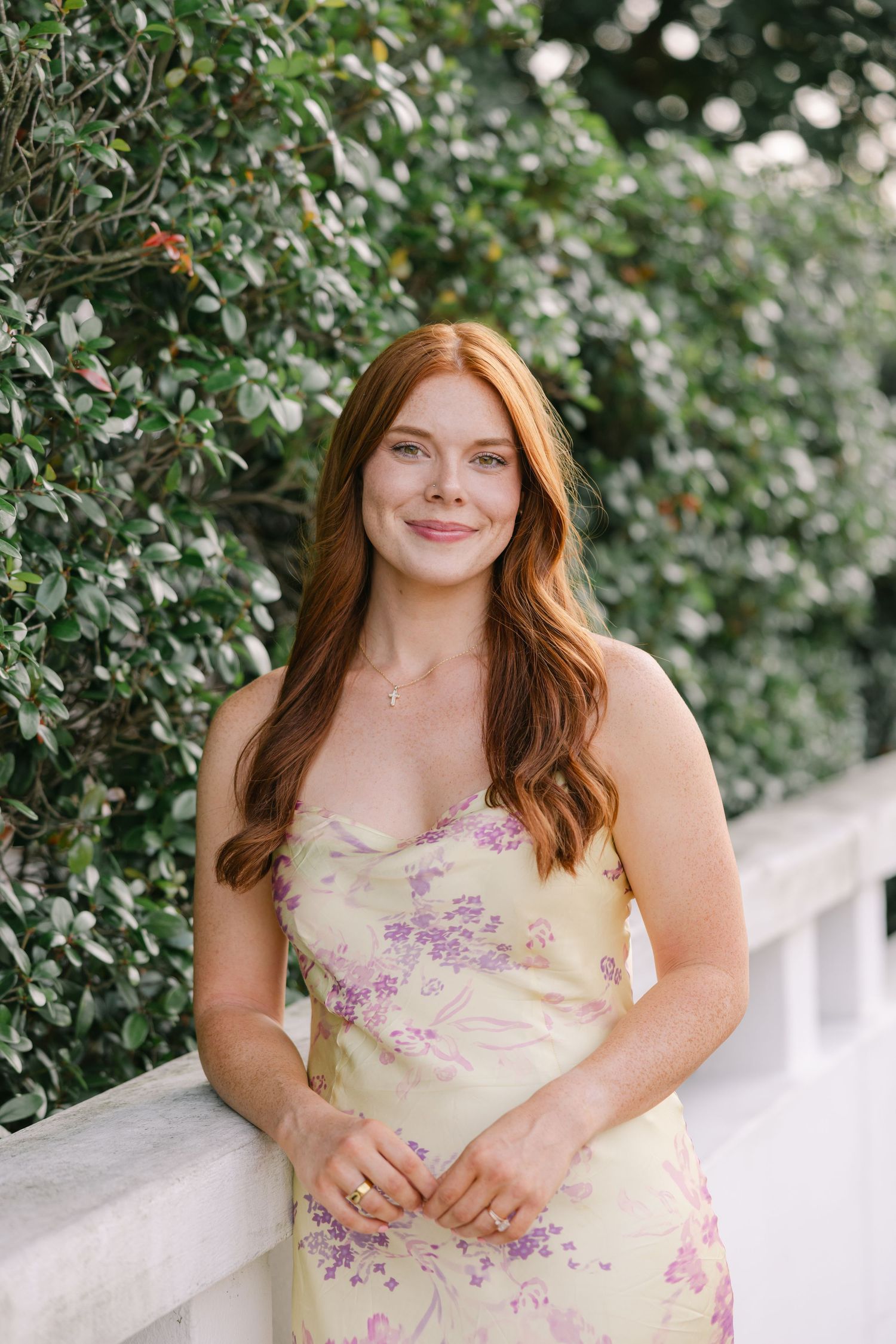 A young red-headed woman smiles softly at the camera in front of beautiful greenery in soft lighting. LSU GRAD PHOTOGRAPHER