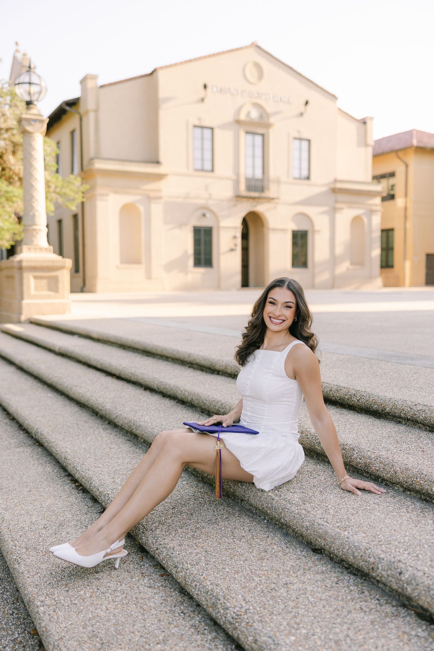 A young college graduate beams at the camera, sitting on stone architectural steps posing with her graduation cap. LSU GRAD PHOTOGRAPHER