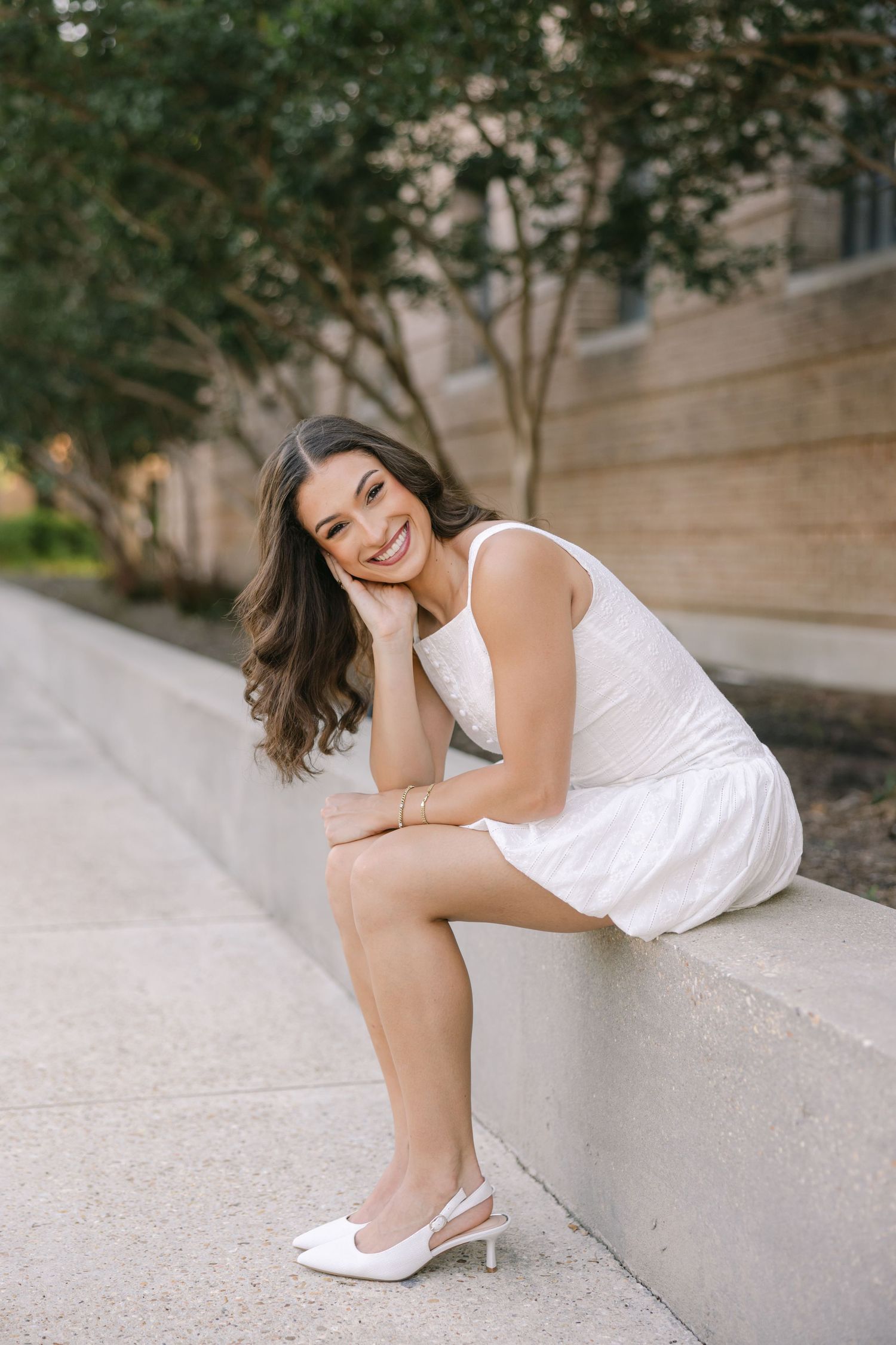 A young woman leans on her knees and smiles at the camera in front of a stone and tree lined pathway