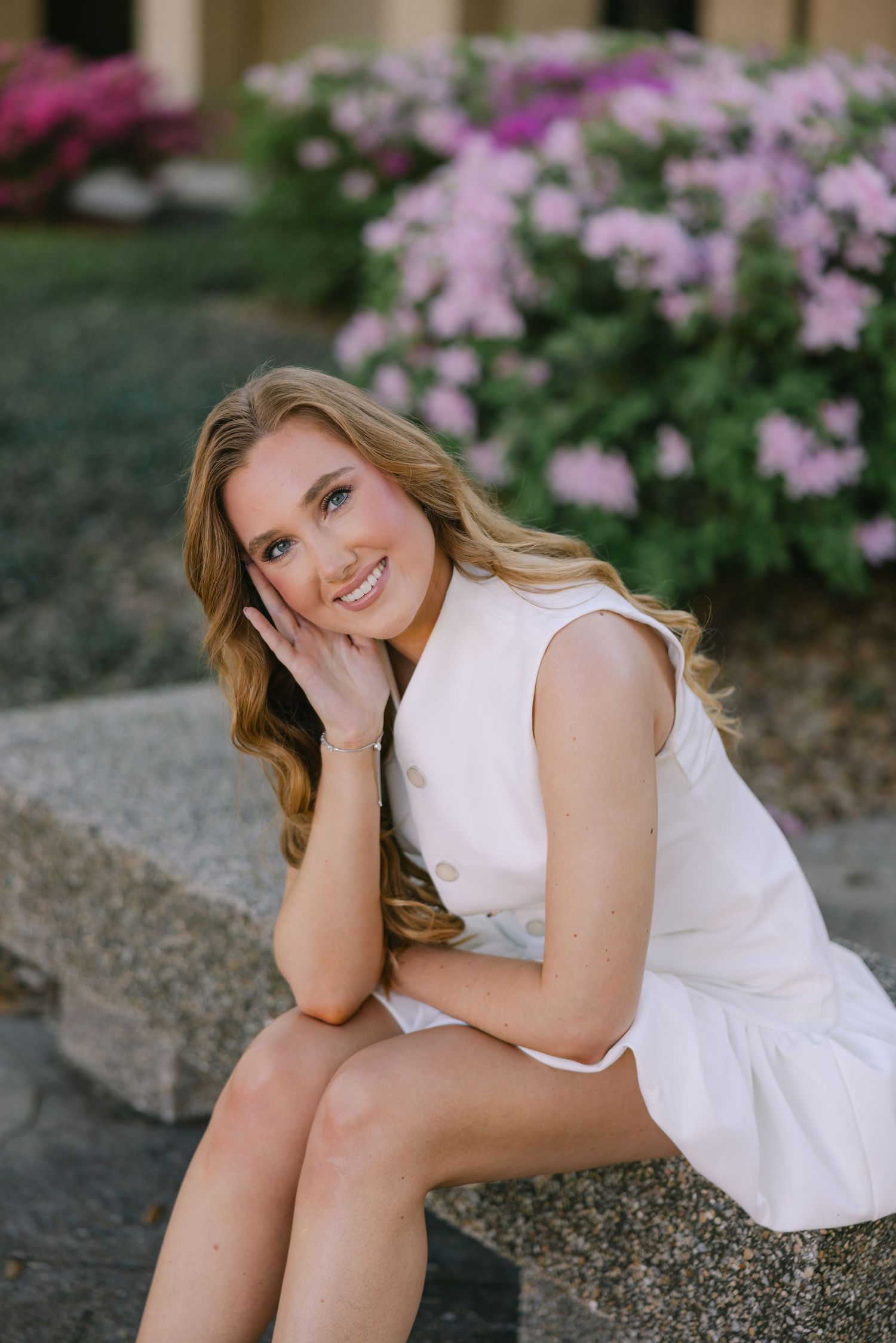 A young woman leans on her knees and smiles at the camera in front of a flower bush